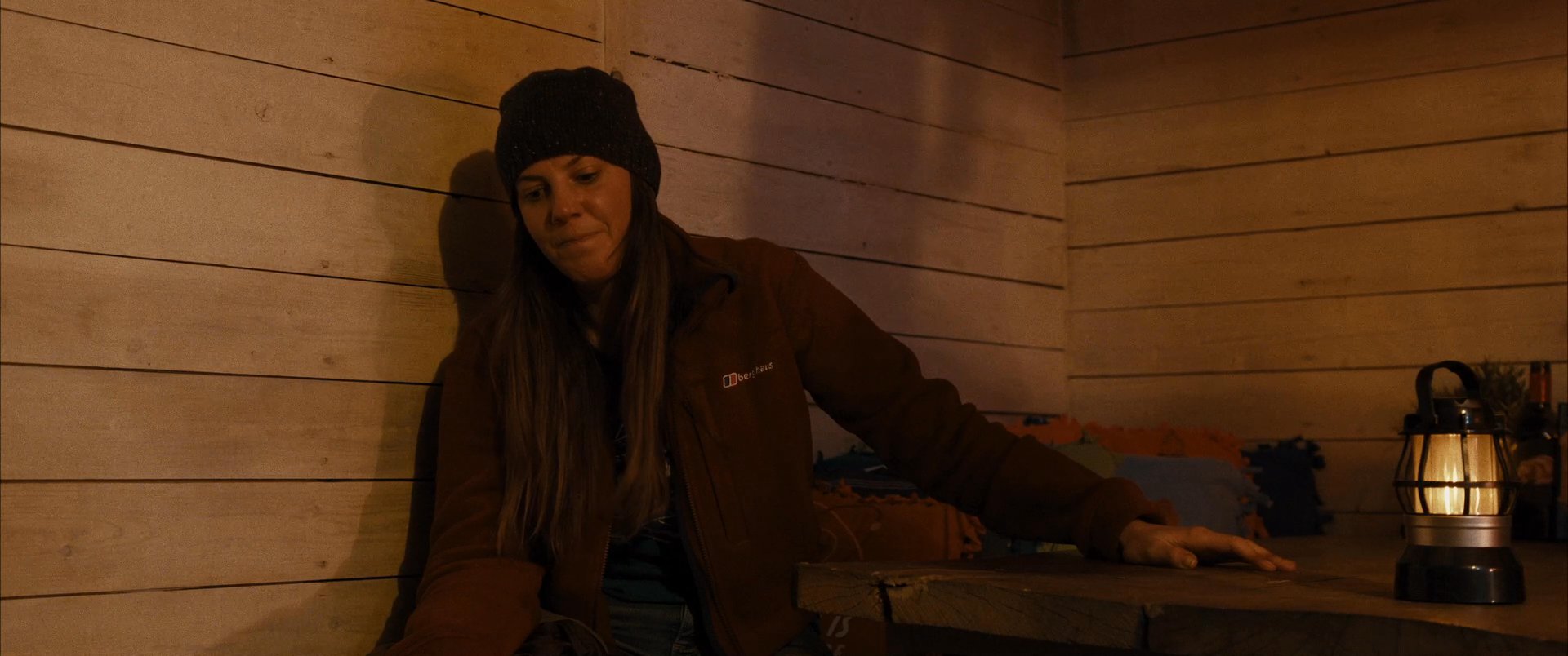 Two individuals engage in a somber, intimate conversation within a dimly lit, rustic cabin setting. They alternate speaking while seated at a table illuminated by a lantern, suggesting a narrative exchange.