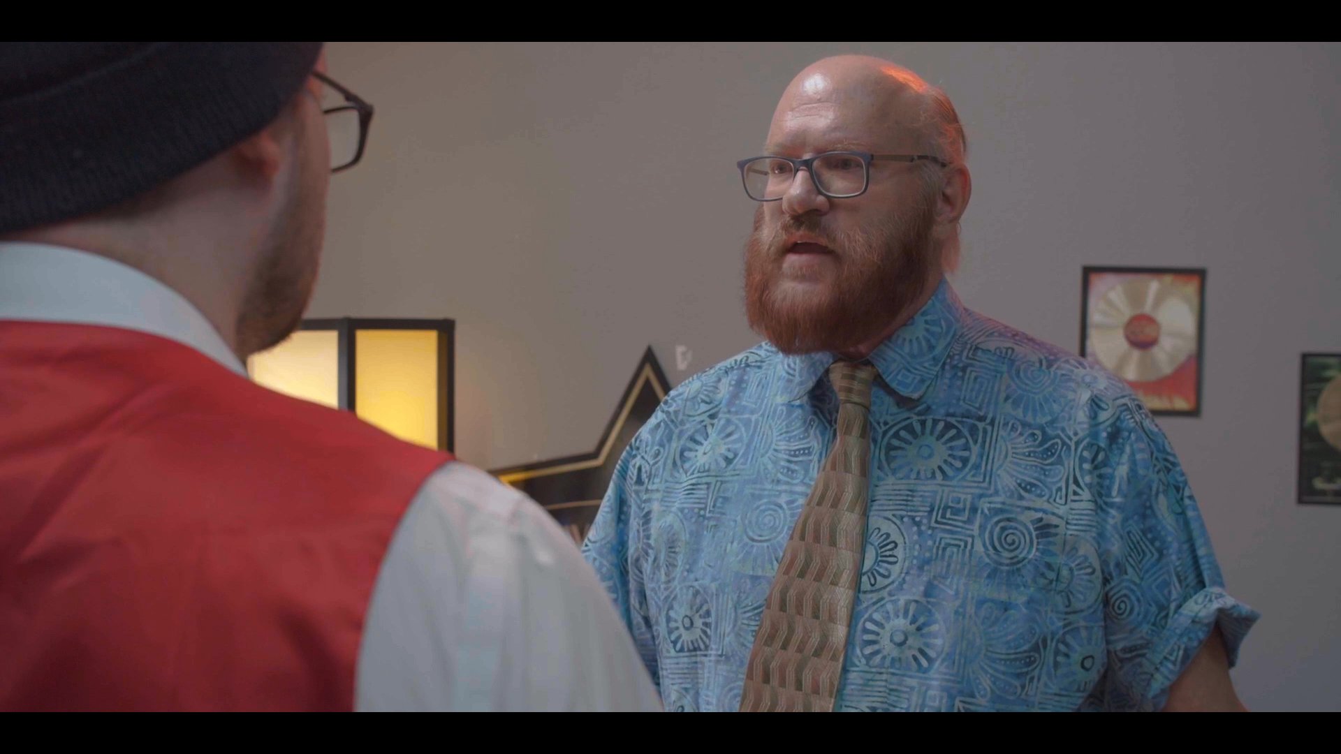 Two colleagues engage in a tense, animated discussion across an office desk, debating terms while referencing small items arranged between them.