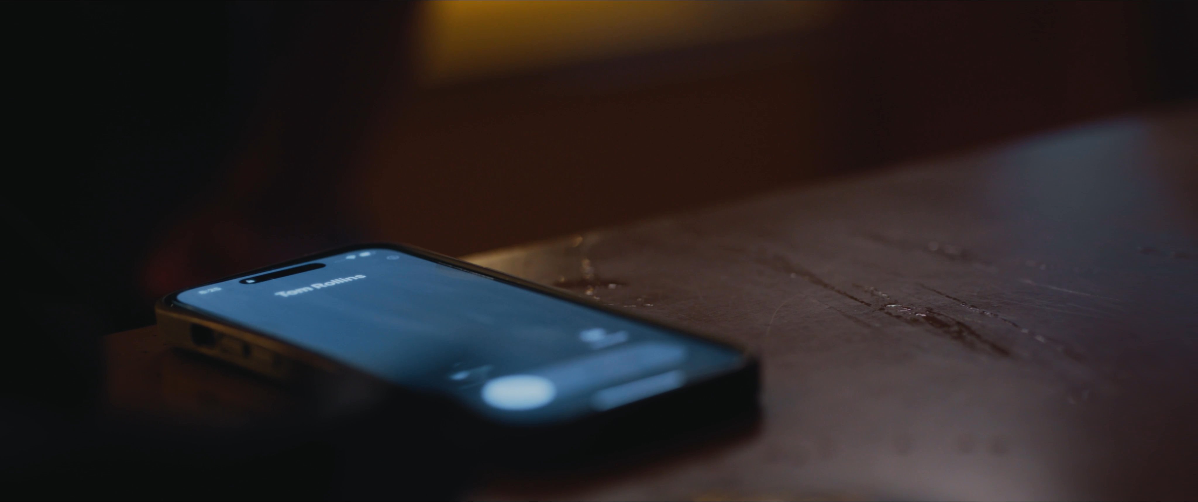 A man in a dim kitchen notices an incoming call on his smartphone resting on a counter. He retrieves the device and prepares to answer it while standing in the quiet room.