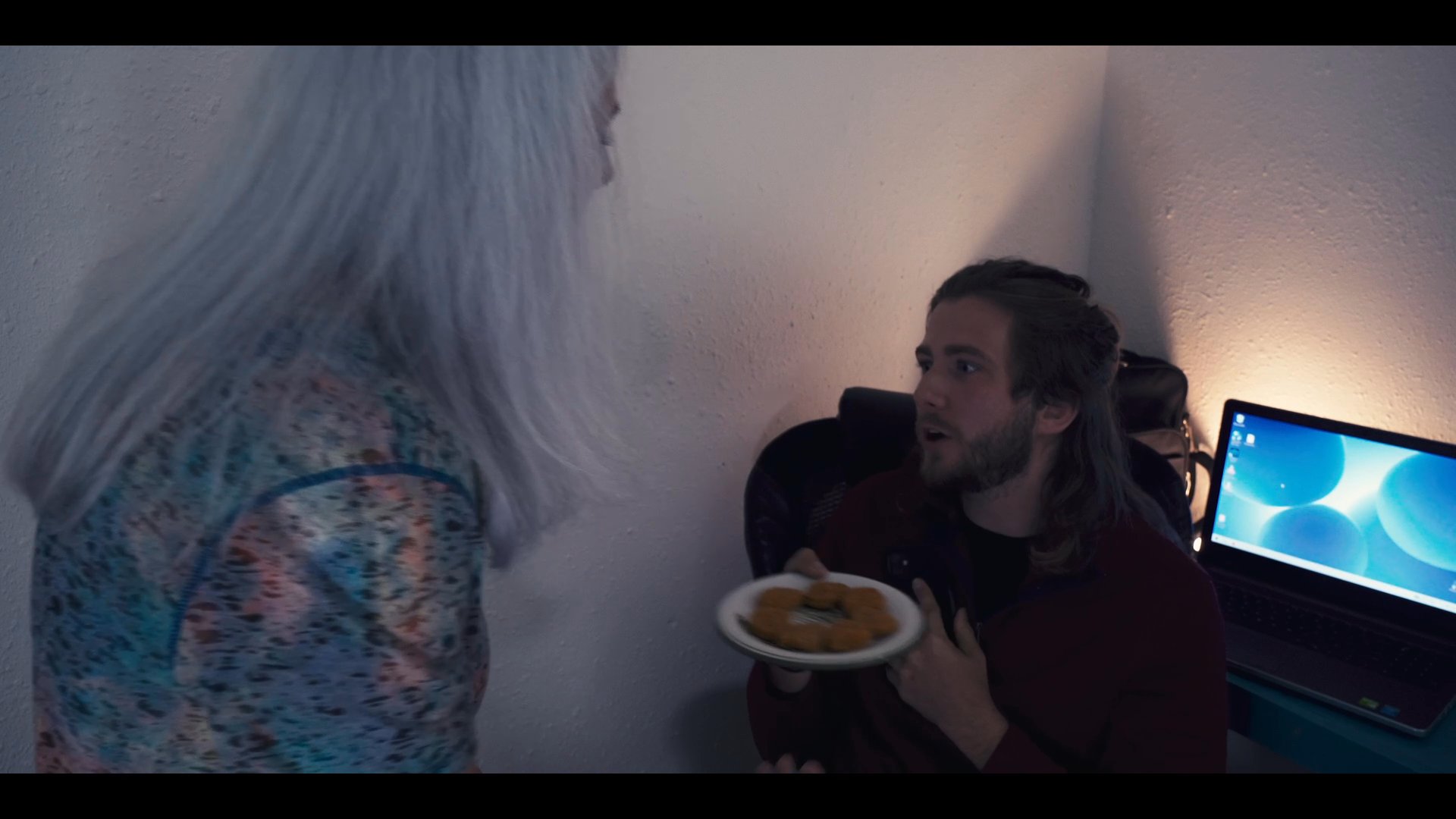 A man manages a phone conversation while transitioning between a bedroom and a home office space, where he is interrupted by his partner offering food. The scene captures the intersection of personal communication and routine home life.