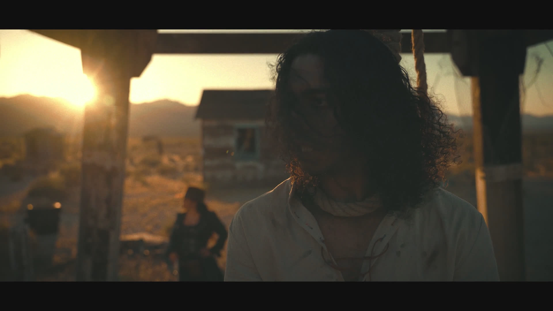 At sunset in a dusty frontier settlement, a dark-clad figure in a wide-brimmed hat confronts a long-haired man in a white shirt. Their tense exchange unfolds beside weathered buildings.