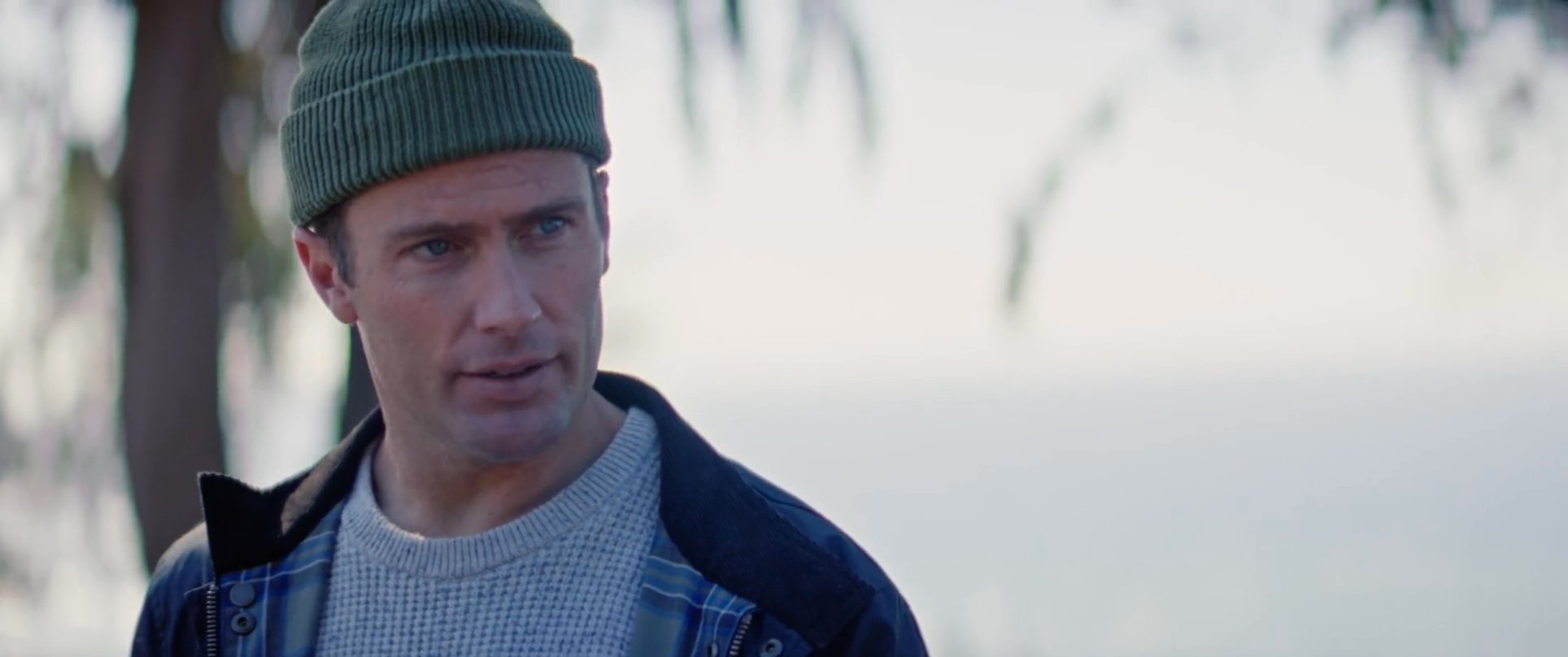 Two men wearing beanies and sunglasses sit on a bench near the coast, engaging in a focused conversation while drinking from metal mugs.