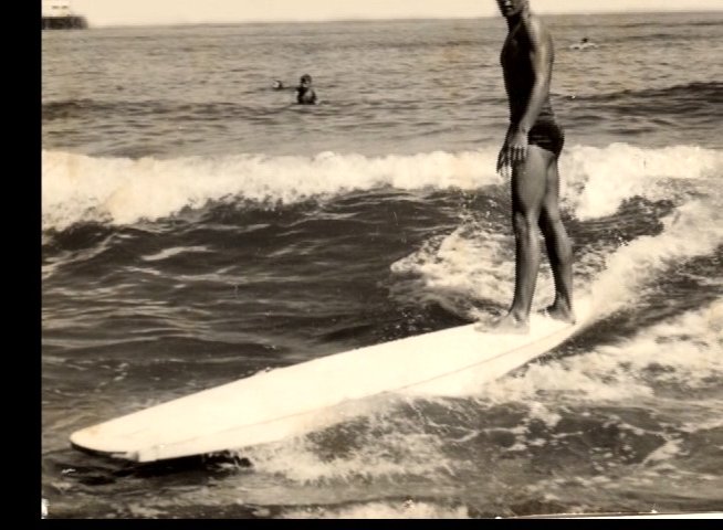 An experienced surfer reflects on early surfing experiences in Santa Cruz, illustrated by archival footage of him riding waves on a longboard.