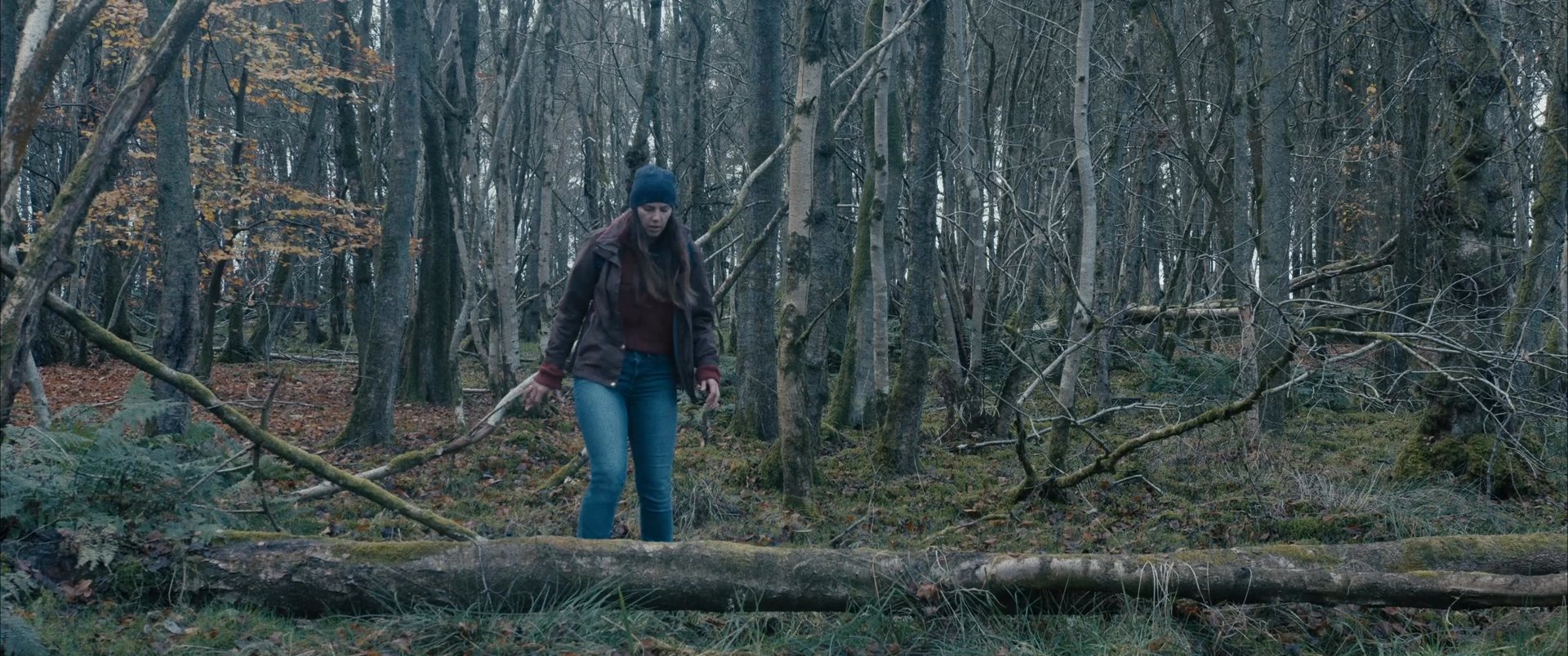 A woman walks through a dense, rustic woodland area, navigating uneven terrain while surrounded by tall, leafless trees.