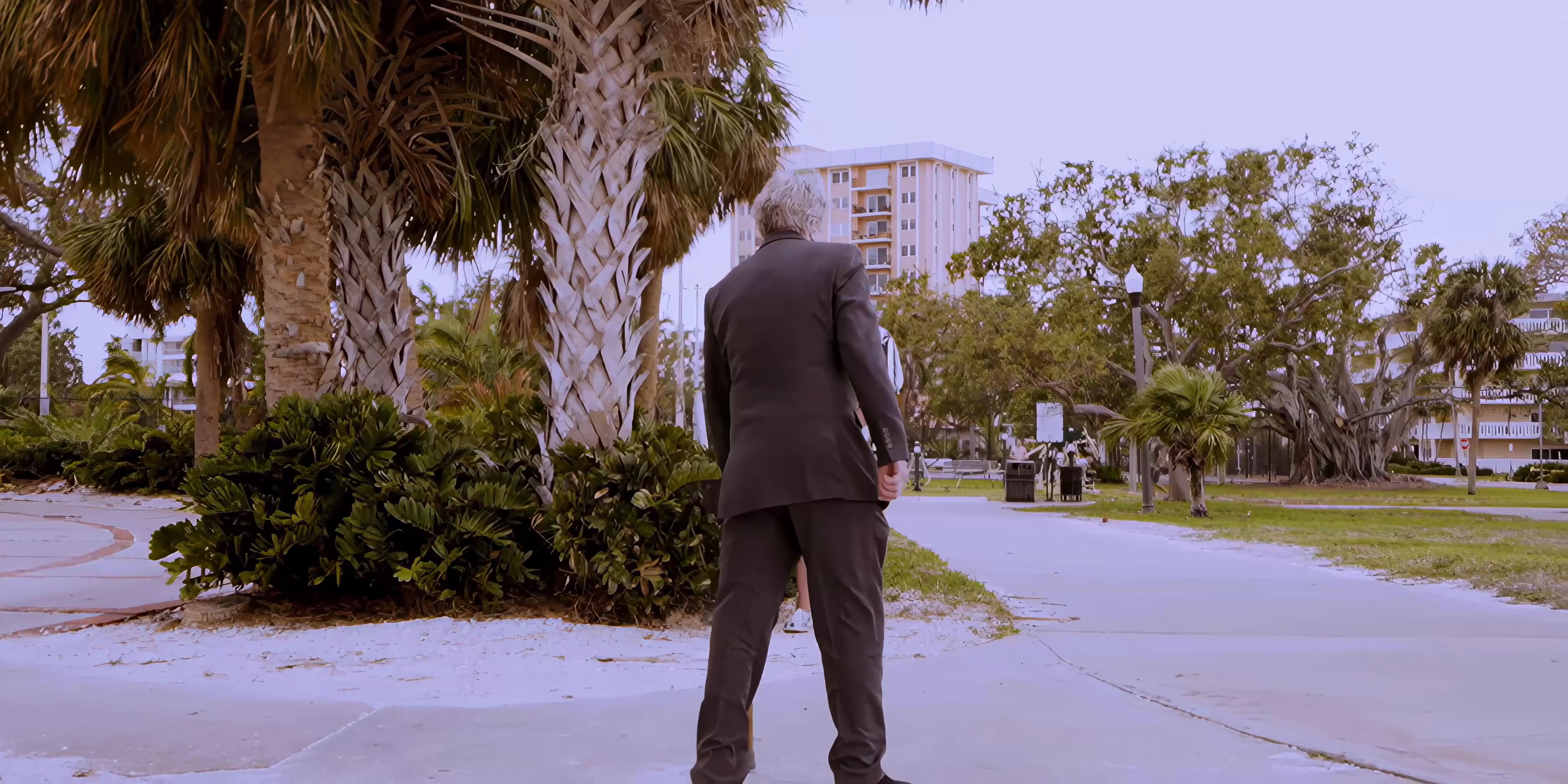 An older man in a suit engages in a brief interaction in a public park before transitioning to a scenic city aerial view and a reflective car ride.