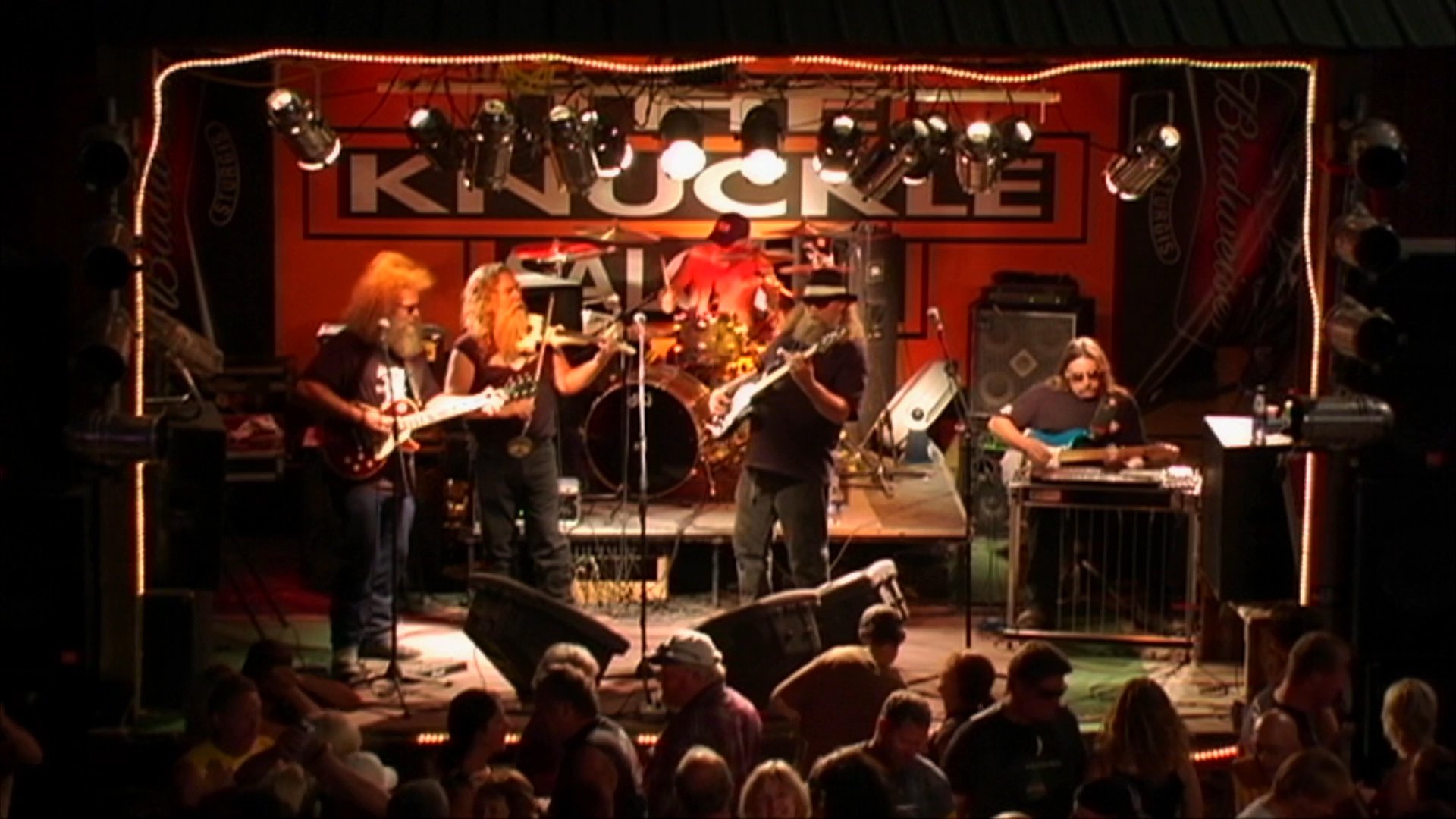 A band performs live on stage at a concert venue, featuring a violinist and guitarist leading the ensemble. The scene captures the high-energy musical interaction between the performers and the band's instrumental variety.