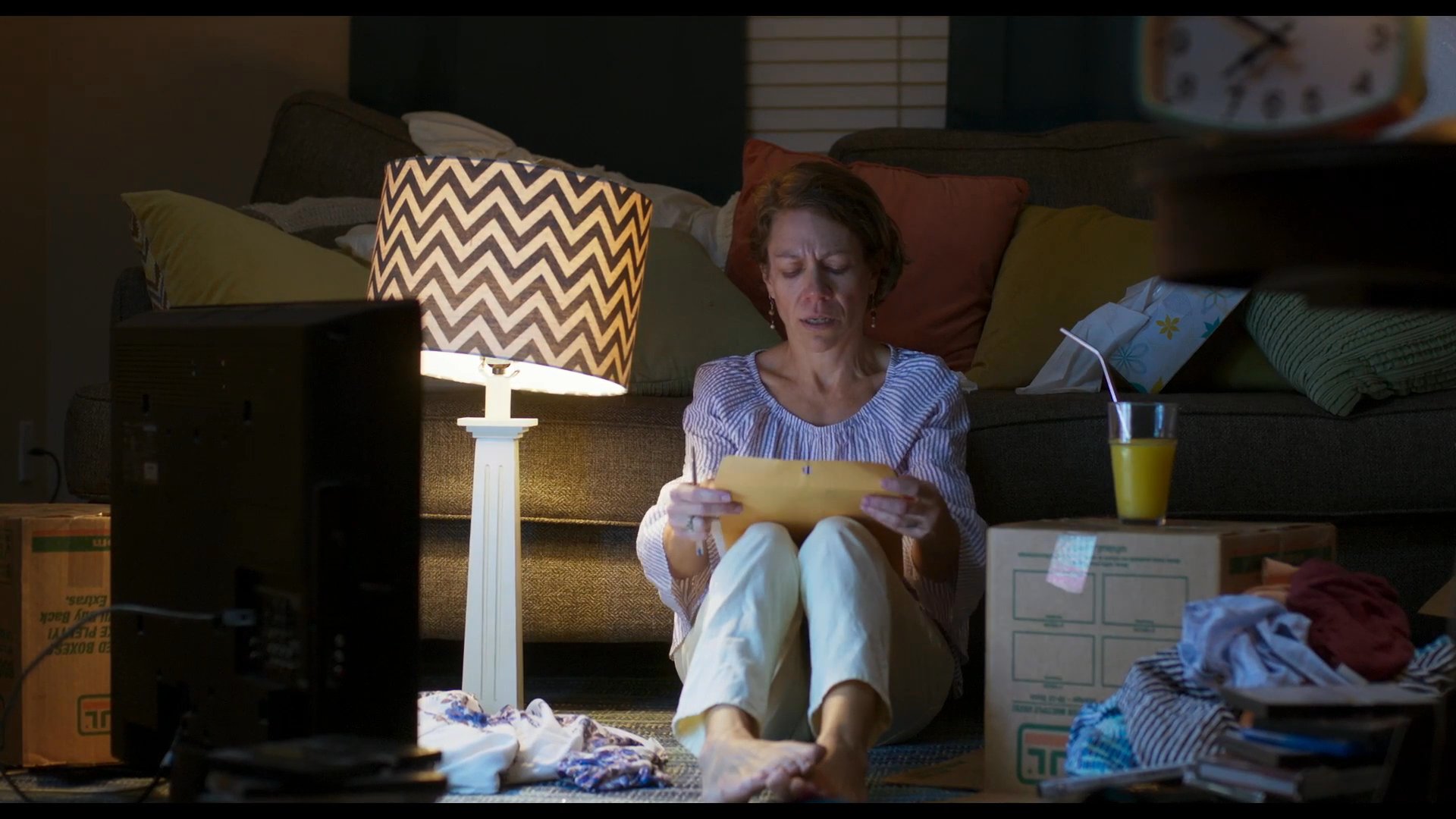 A woman sits on the floor of a dimly lit room, surrounded by moving boxes, while carefully reading through documents. She appears pensive and focused as she processes the information in a quiet home environment.