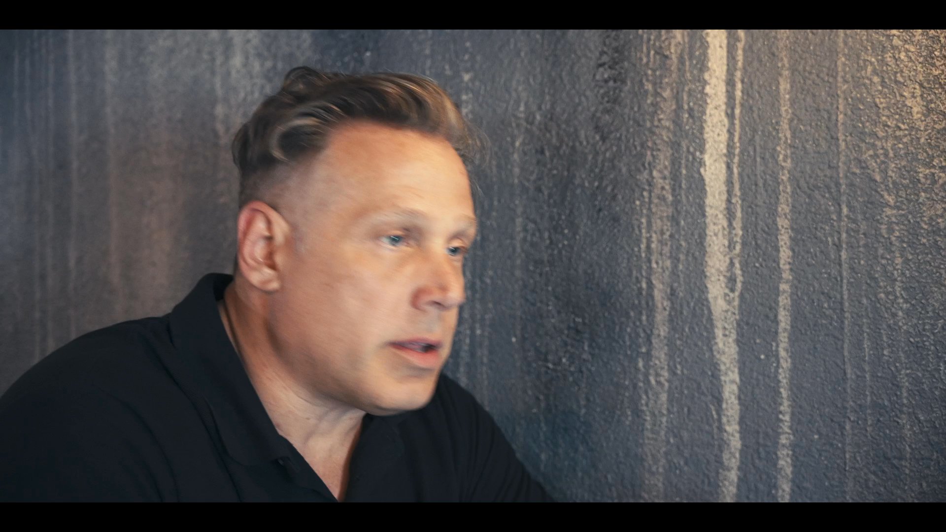 Two individuals engage in a focused, seated conversation against a textured backdrop, alternating between speaking and listening roles.