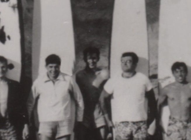 A group of surfers stands together on a sandy beach, posing alongside their surfboards in a vintage black and white photograph.