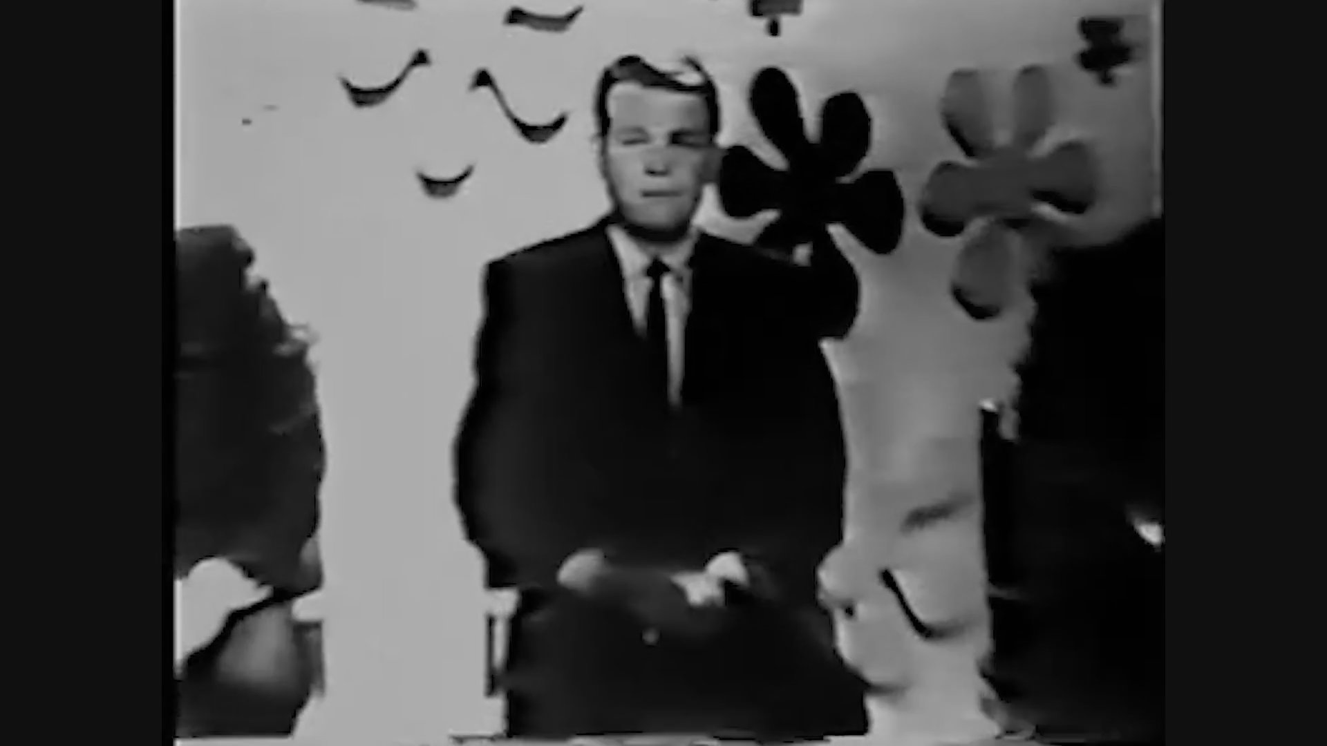 A charismatic television host introduces panelists on the set of a classic game show. The scene transitions between wide shots of the stage and close-ups of the participants in a studio environment.