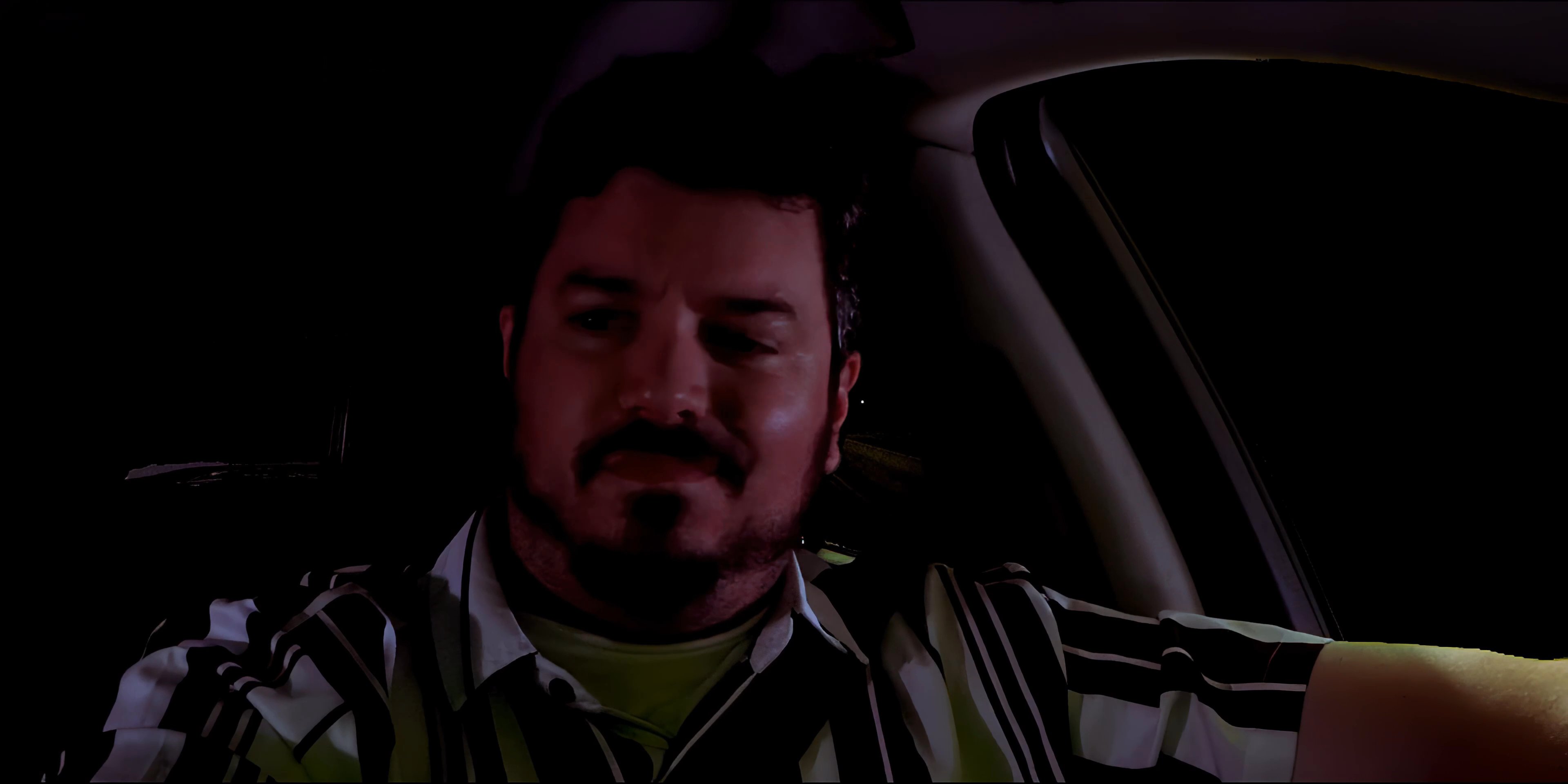 A man sits in the driver's seat of a car at night, engaging in conversation while traveling along an illuminated street.