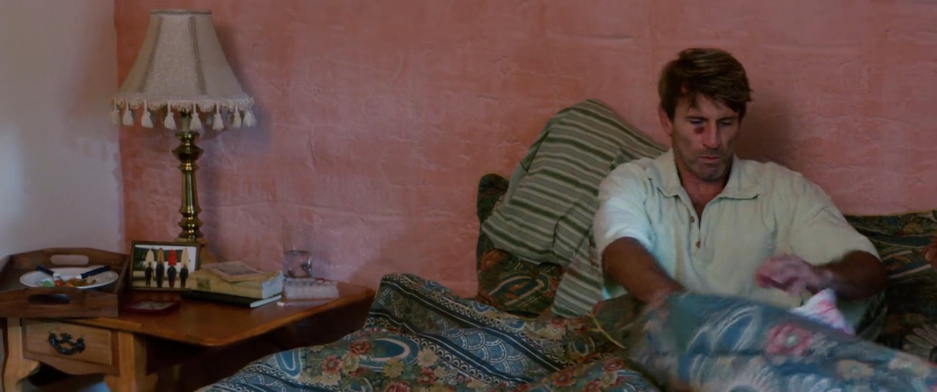 A man wakes up in bed to take medication while visitors stand in the doorway of his room. The interaction transitions from a quiet morning routine to a conversation with the men arriving at the bedside.