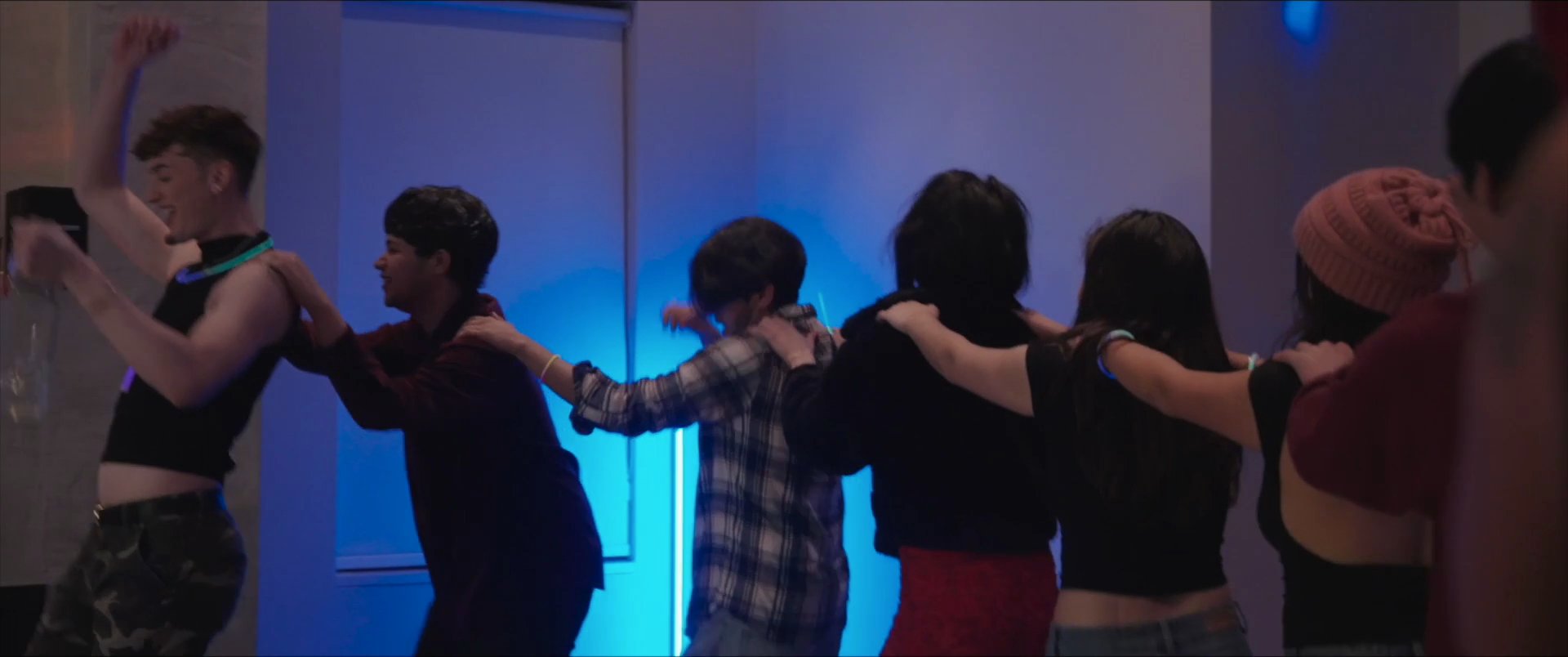 A group of young adults participates in a lively dance at an indoor social gathering. The scene features friends socializing, forming a conga line, and enjoying festive lighting.