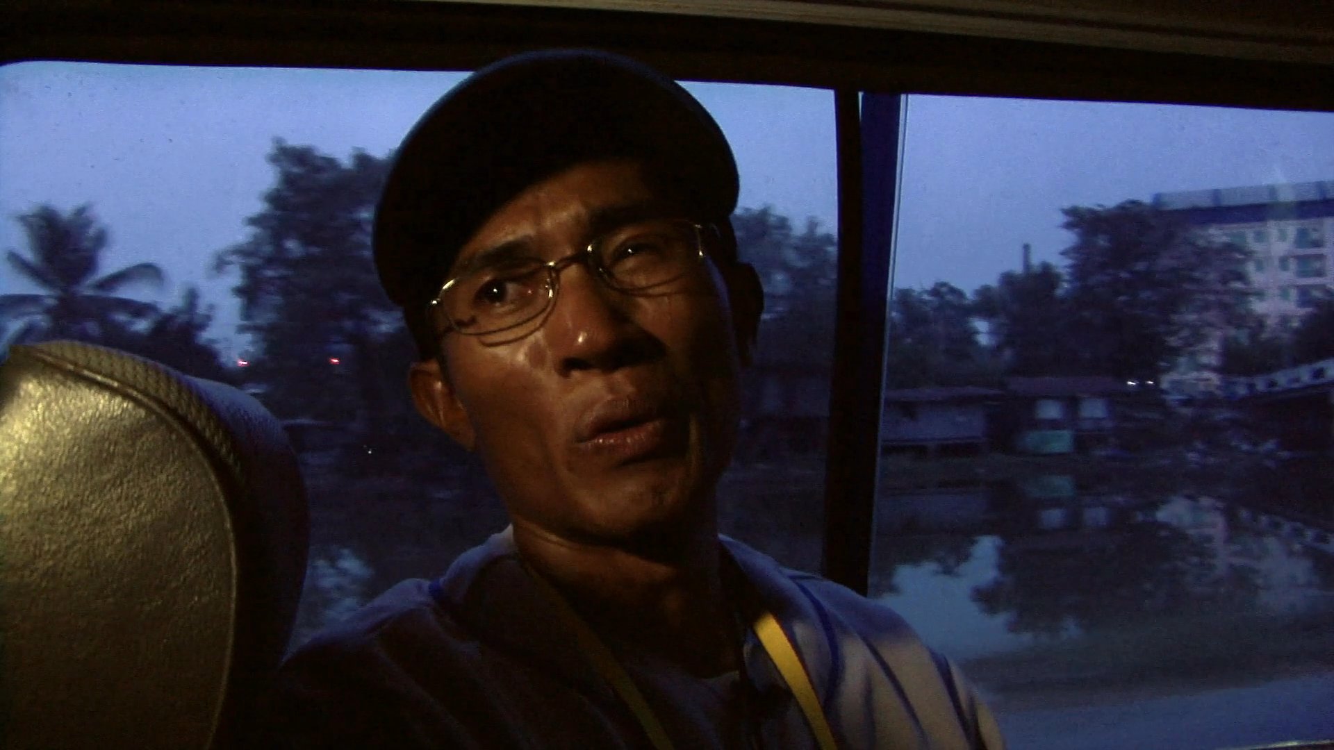 A man wearing a baseball cap speaks while riding as a passenger in a vehicle driving through a city during twilight.