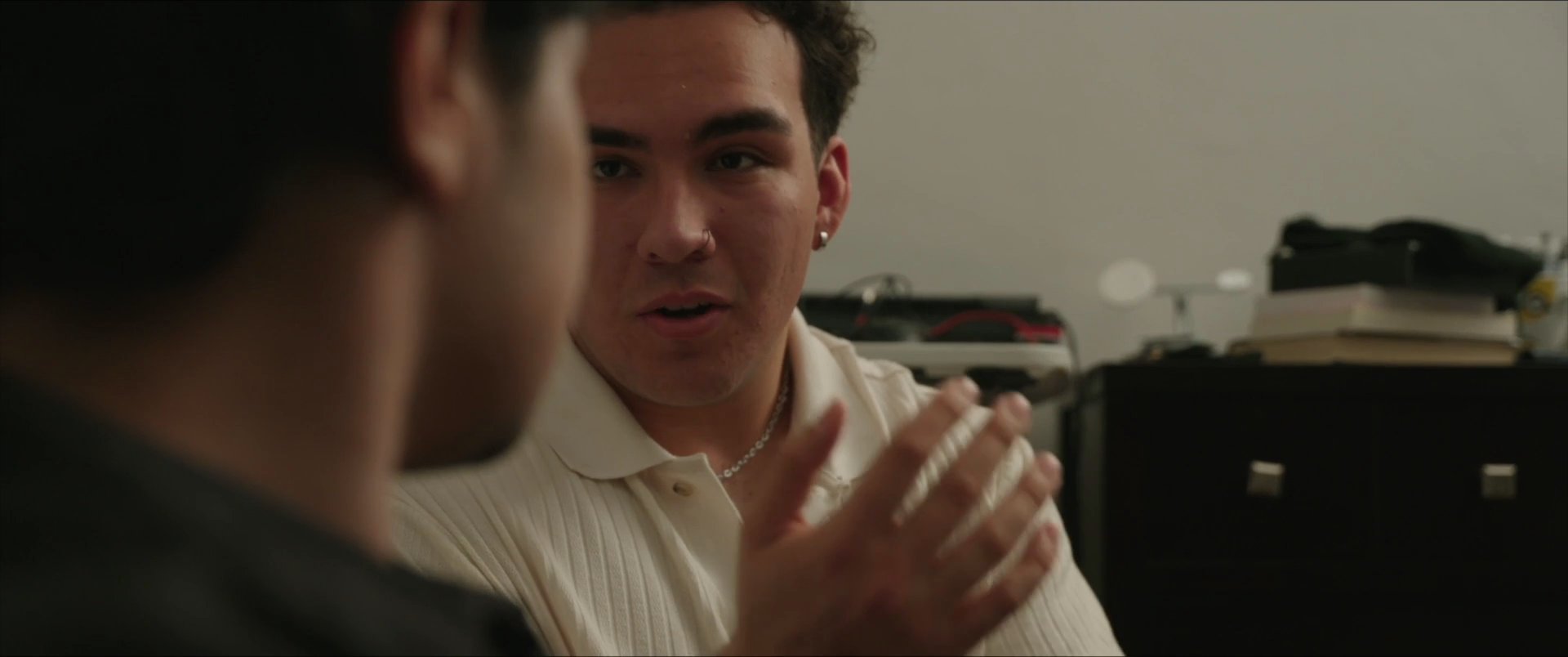 Two young men engage in a focused, face-to-face conversation within an indoor residential setting. Their interaction centers on verbal exchange and sustained eye contact.