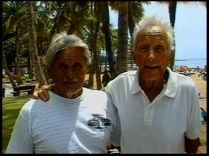 Two older men engage in a friendly conversation on a sunny beach before transitioning to a scenic drive along the coast in a vintage truck. The sequence captures a shared moment of camaraderie and island exploration.
