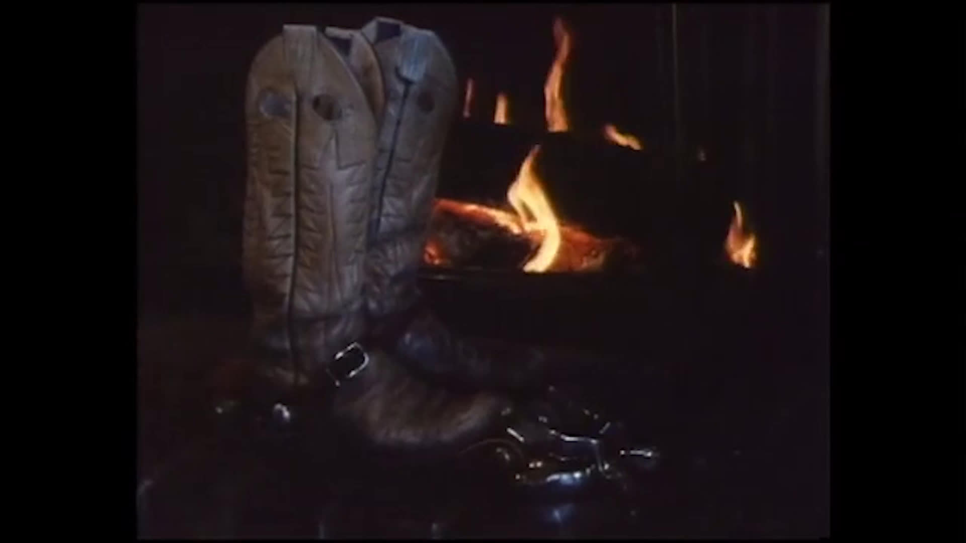 Snowy lakeshores and a pine-lined cabin set a quiet winter scene. Inside, cowboy boots stand beside a glowing fireplace as a man and two children gather; the accompanying song celebrates cowboys and the rodeo.