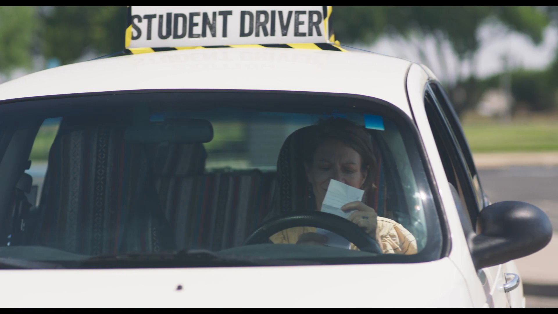 An instructor waits in a student driver vehicle outside a school before picking up a student who approaches the car to begin a driving lesson.