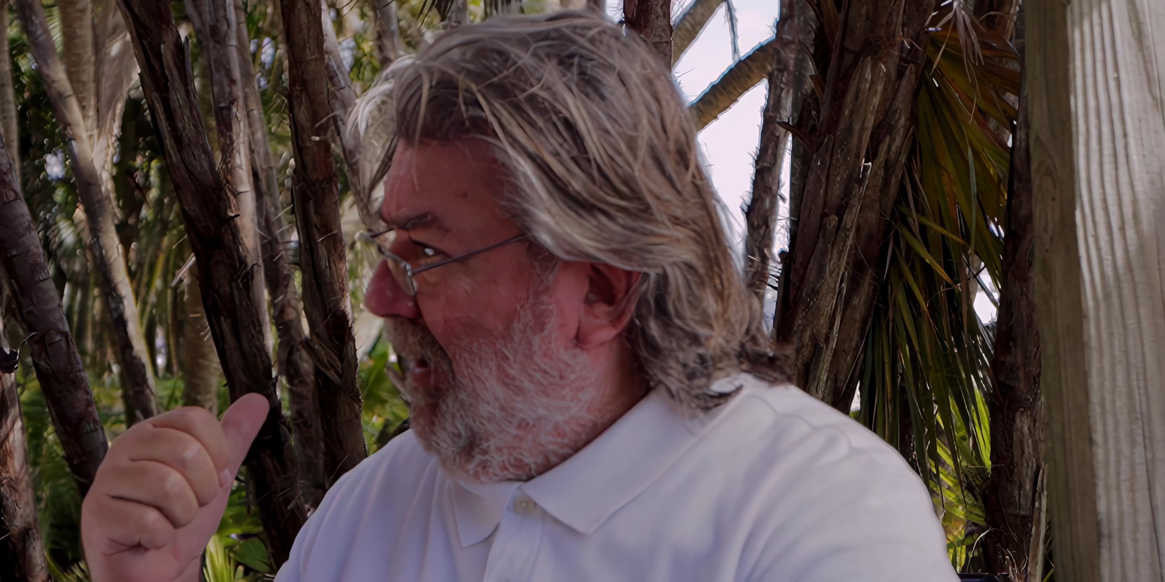 A man with a gray beard sits outdoors and engages in a thoughtful conversation while gesturing to emphasize his points. Another man in a suit listens attentively while seated nearby.