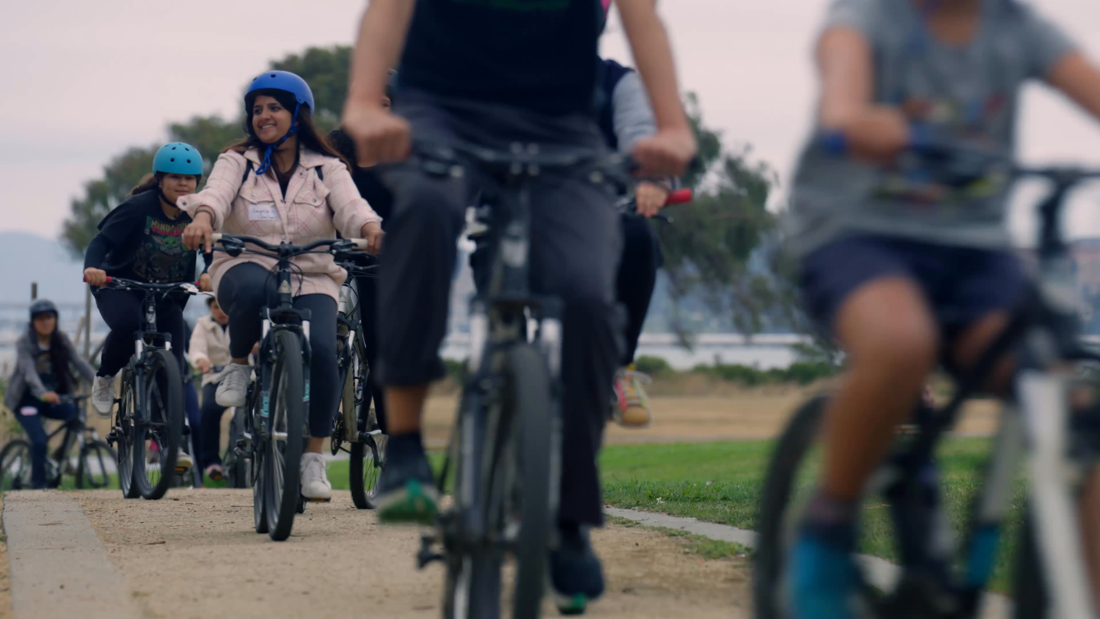 At a community park, a speaker connects protecting local green spaces with public health as volunteers help children with bicycle helmets. Participants ride, run, and use binoculars outdoors, sharing an energetic day of recreation and nature observation.