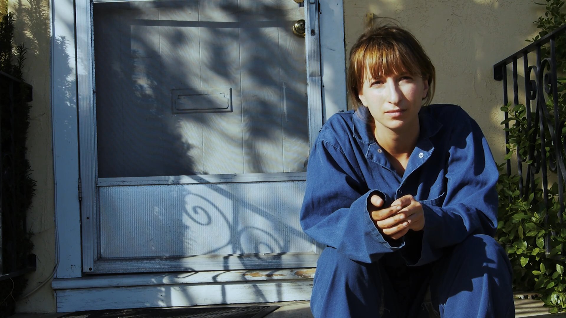 A young man sits behind the steering wheel of a car, transitioning his gaze from the road to the camera, while a young woman in blue workwear sits on the steps outdoors looking towards the car.