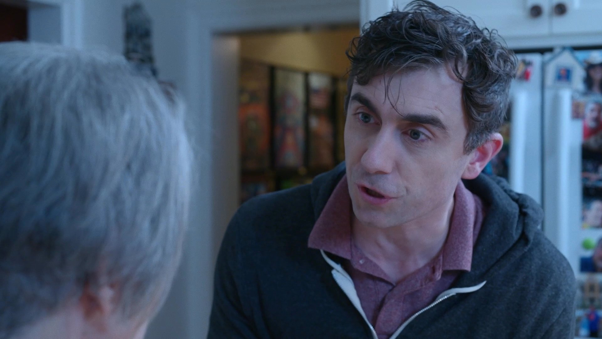 A man and an older woman engage in a serious, focused conversation within a domestic setting. The exchange involves attentive listening and expressive verbal communication between the two individuals.