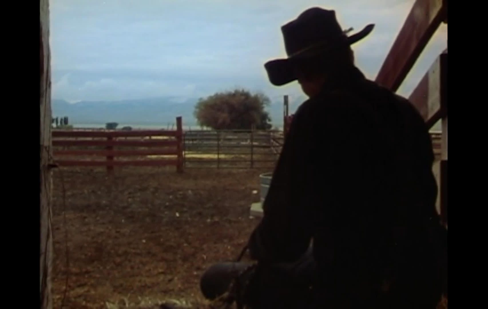 A cowboy in a wide-brimmed hat sits quietly beside a ranch fence, silhouetted against an open pasture. Close-ups of an older man speaking and singing bring a reflective tone, while the sequence opens on a snowy sunset landscape.