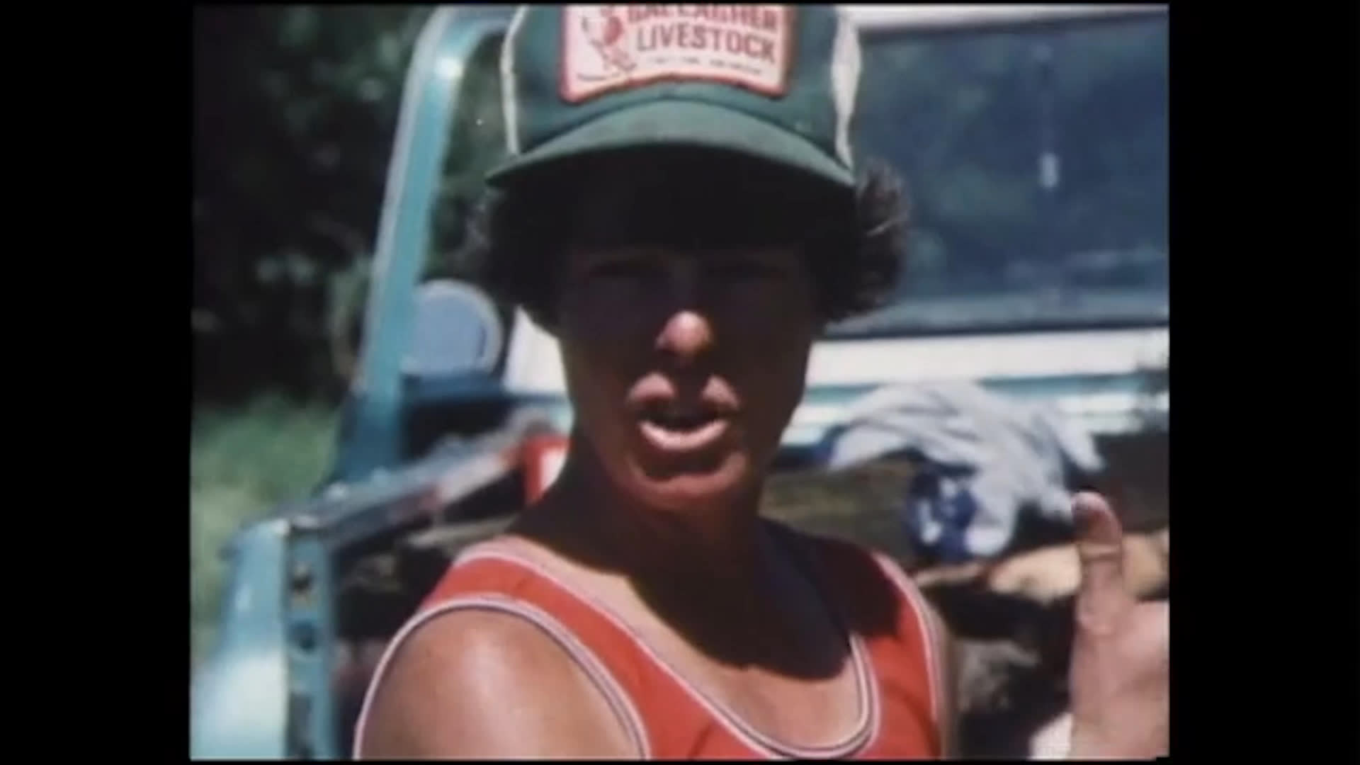 Men gather beside a vehicle outdoors as one recounts an accident while putting gas in a bus. The story ends with a lighthearted family remark and laughter.