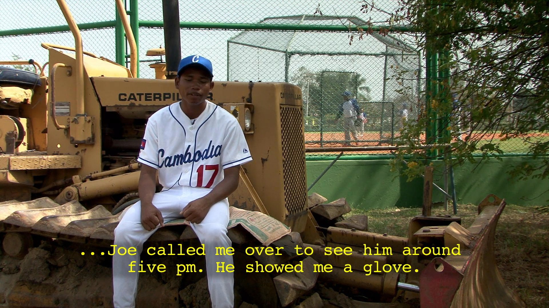 A baseball player in a team uniform discusses his experience being introduced to the sport and learning to use a glove. He sits on heavy machinery at a sports facility while recounting his personal history with the game.