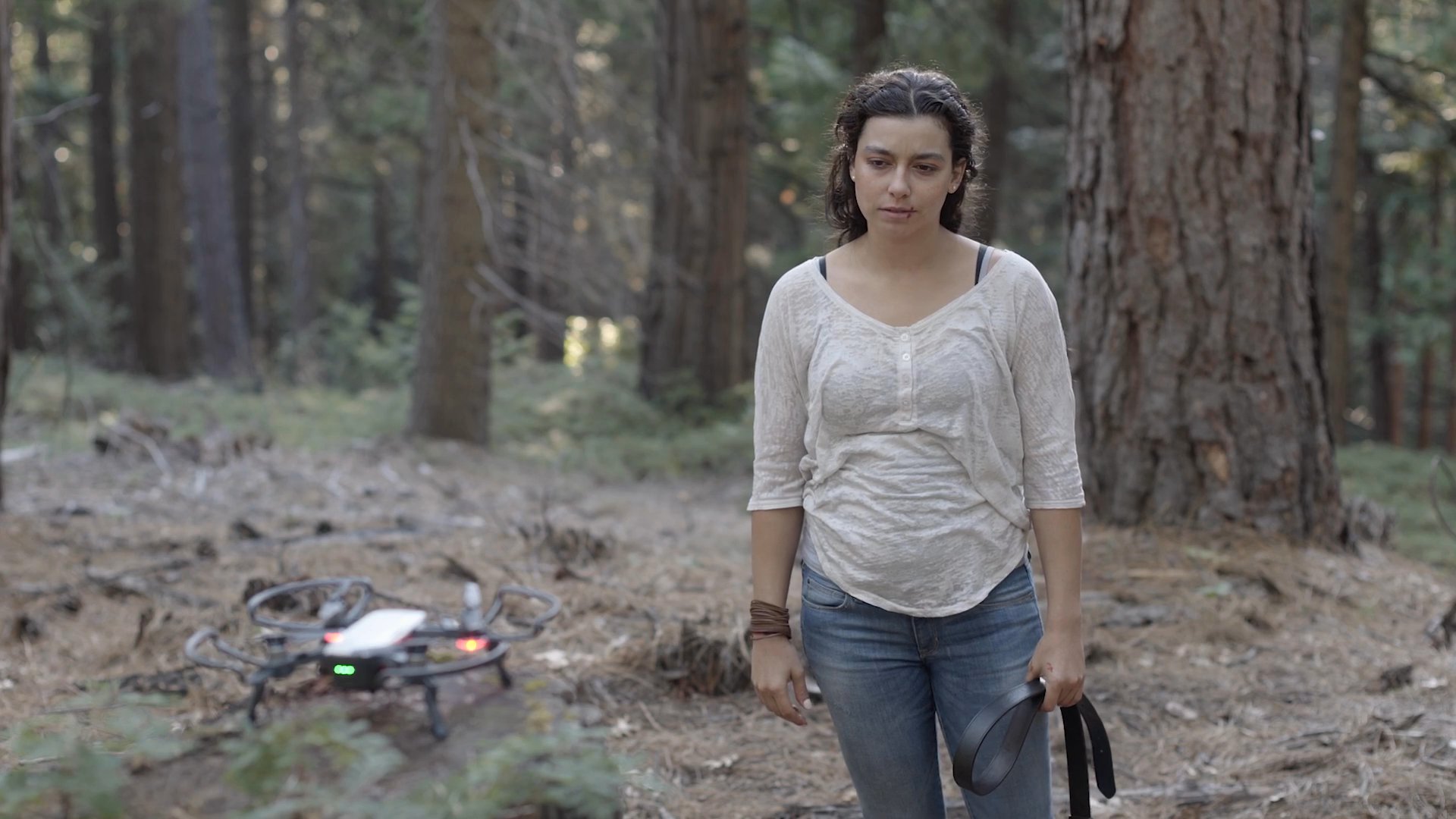 A woman stands in a pine forest, adjusting remote control equipment for a drone resting on a nearby tree stump. She prepares the unmanned aerial vehicle for takeoff before walking away from the device.