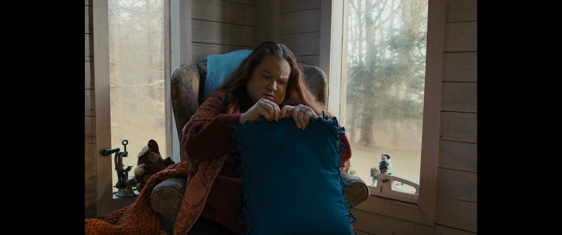A woman sits in an armchair by a window, repeatedly manipulating and clutching a blue cushion with apparent tension. Her repetitive movements suggest a state of restlessness or internal anxiety while in a quiet, indoor setting.