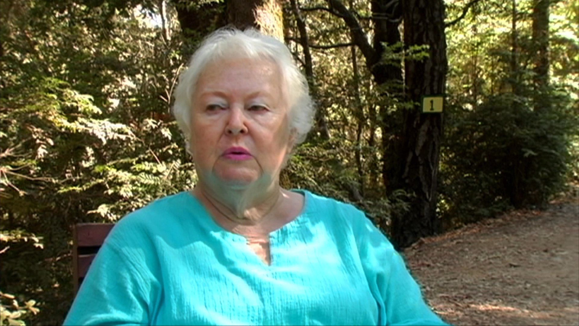 An elderly woman sits in a wooded park setting and shares a personal account while speaking directly to the camera.