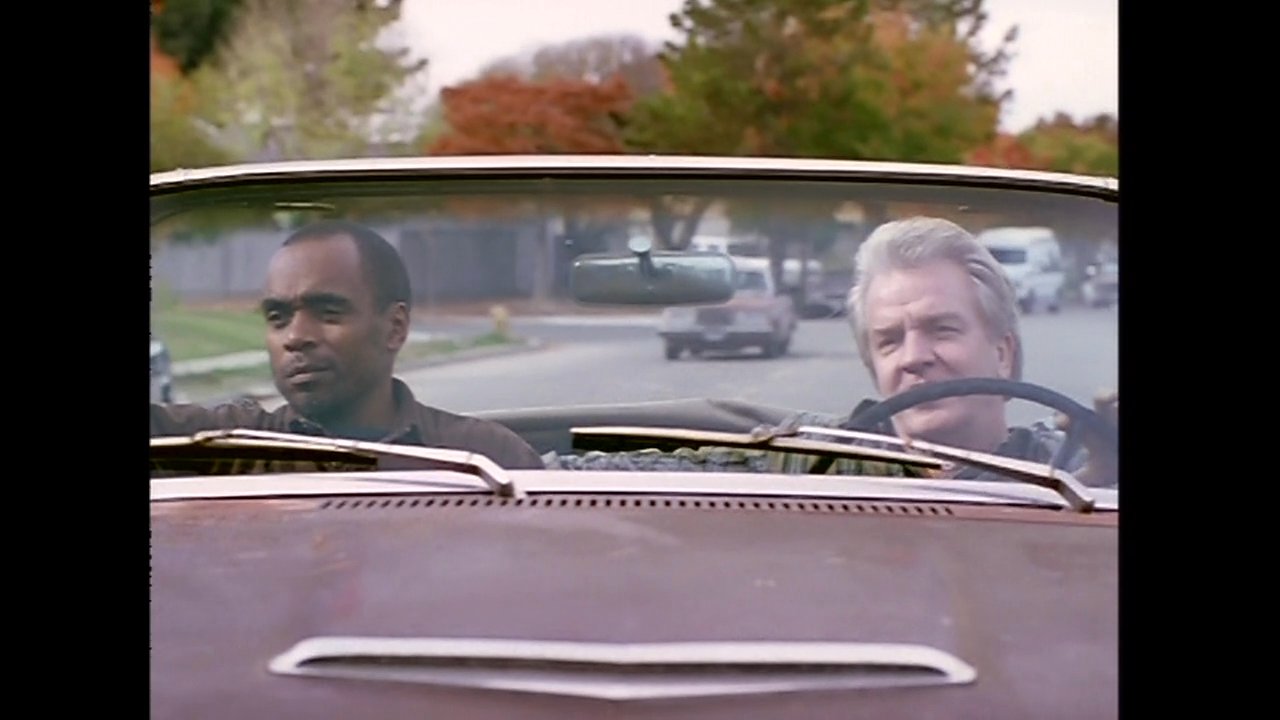 Two men have a conversation while driving through a quiet suburban neighborhood in a vintage convertible car.