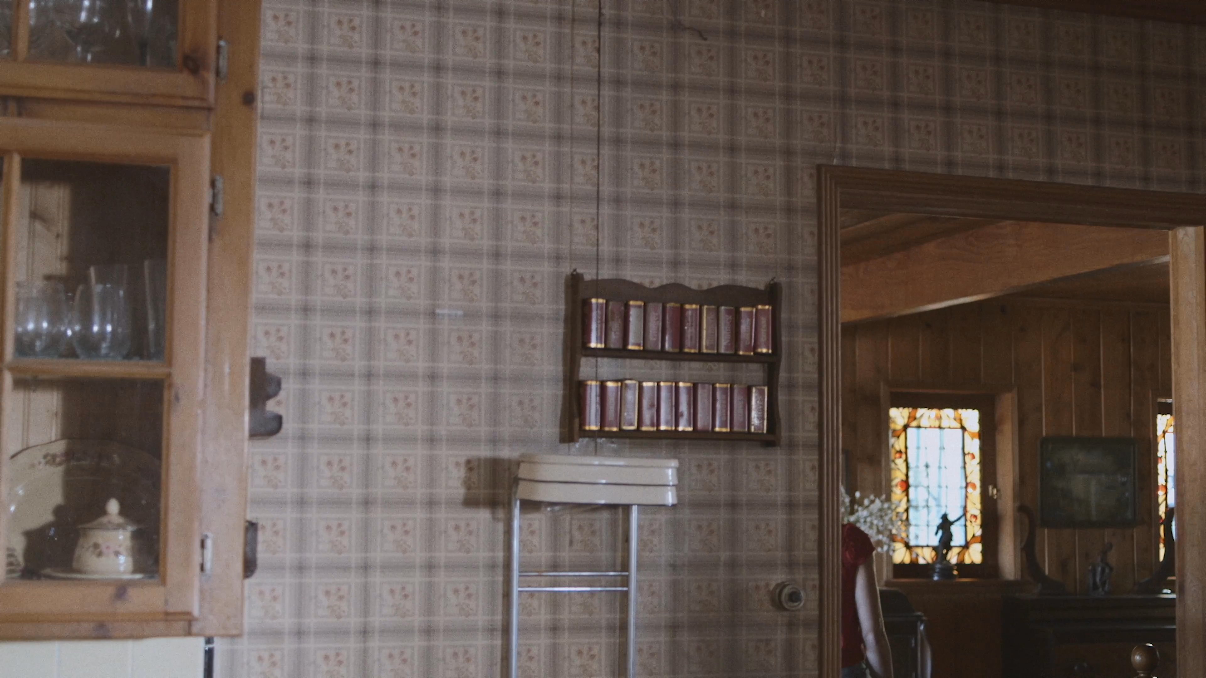 A woman in a red shirt walks through a room and pauses to look out a sunlit window.