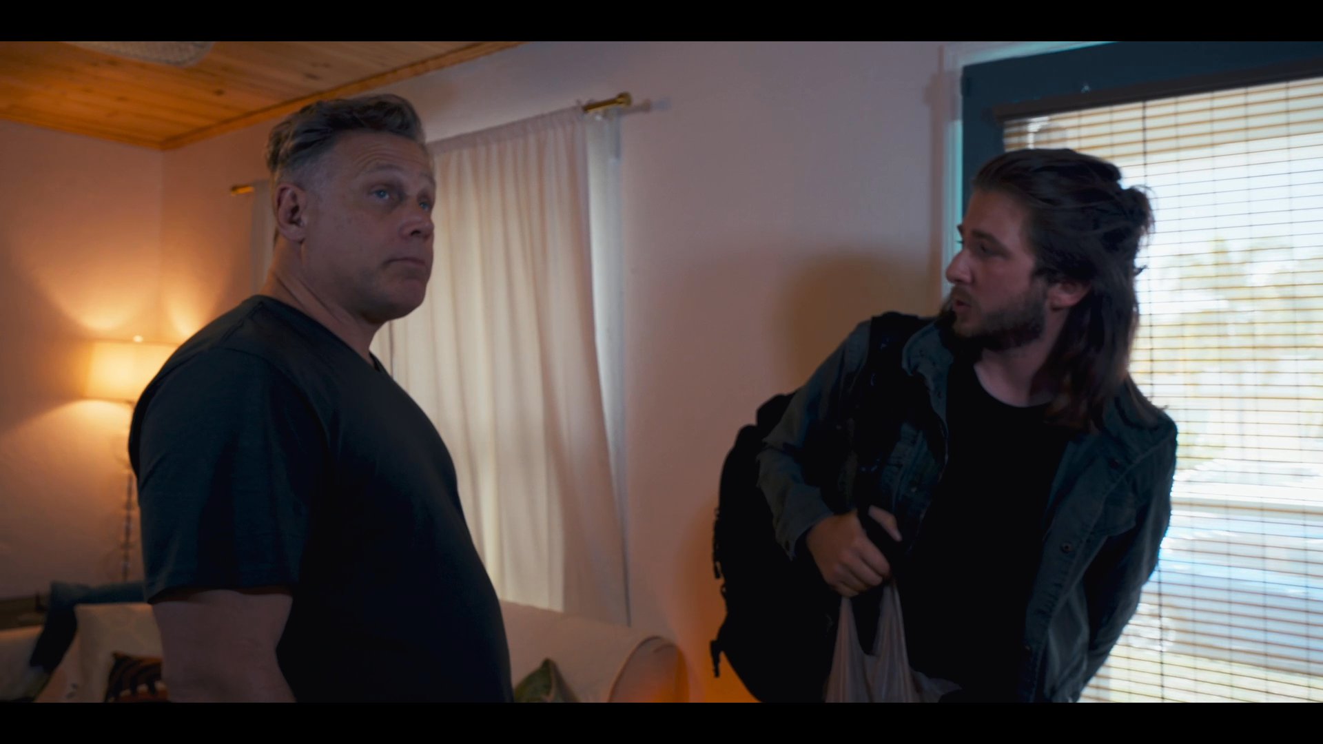 A man opens his front door to welcome a visitor into his home, leading to a brief conversation in the living area. The encounter concludes as they transition toward the kitchen space.