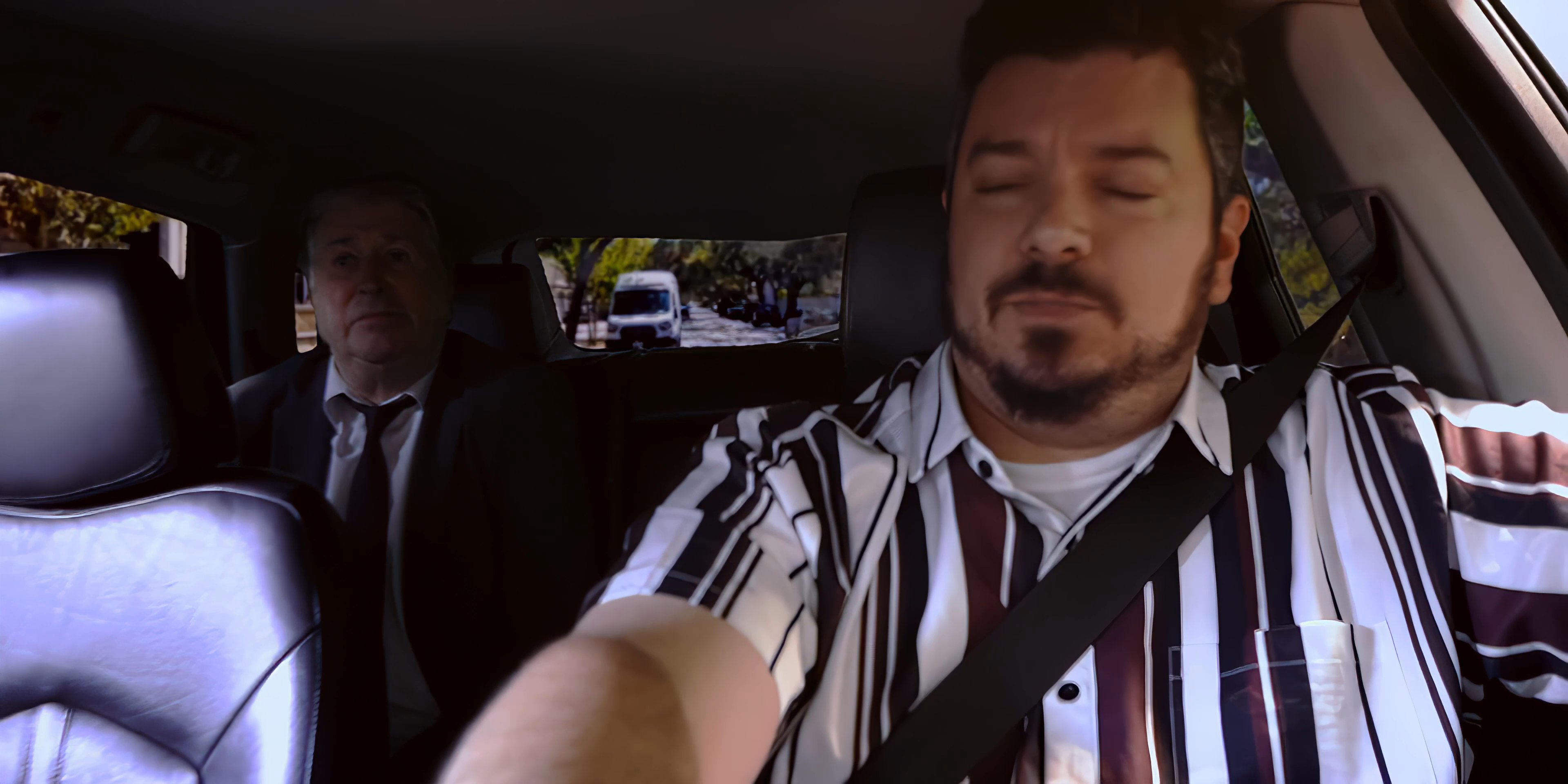 Two individuals engage in a focused discussion while traveling together in the front and back seats of a vehicle. The exterior view shifts to a quiet suburban setting as the conversation continues.