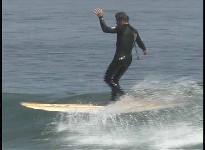 A man in a black wetsuit skillfully maneuvers his surfboard along the crest of a rolling ocean wave. He maintains balance while navigating the breaking water in a calm, open-sea setting.