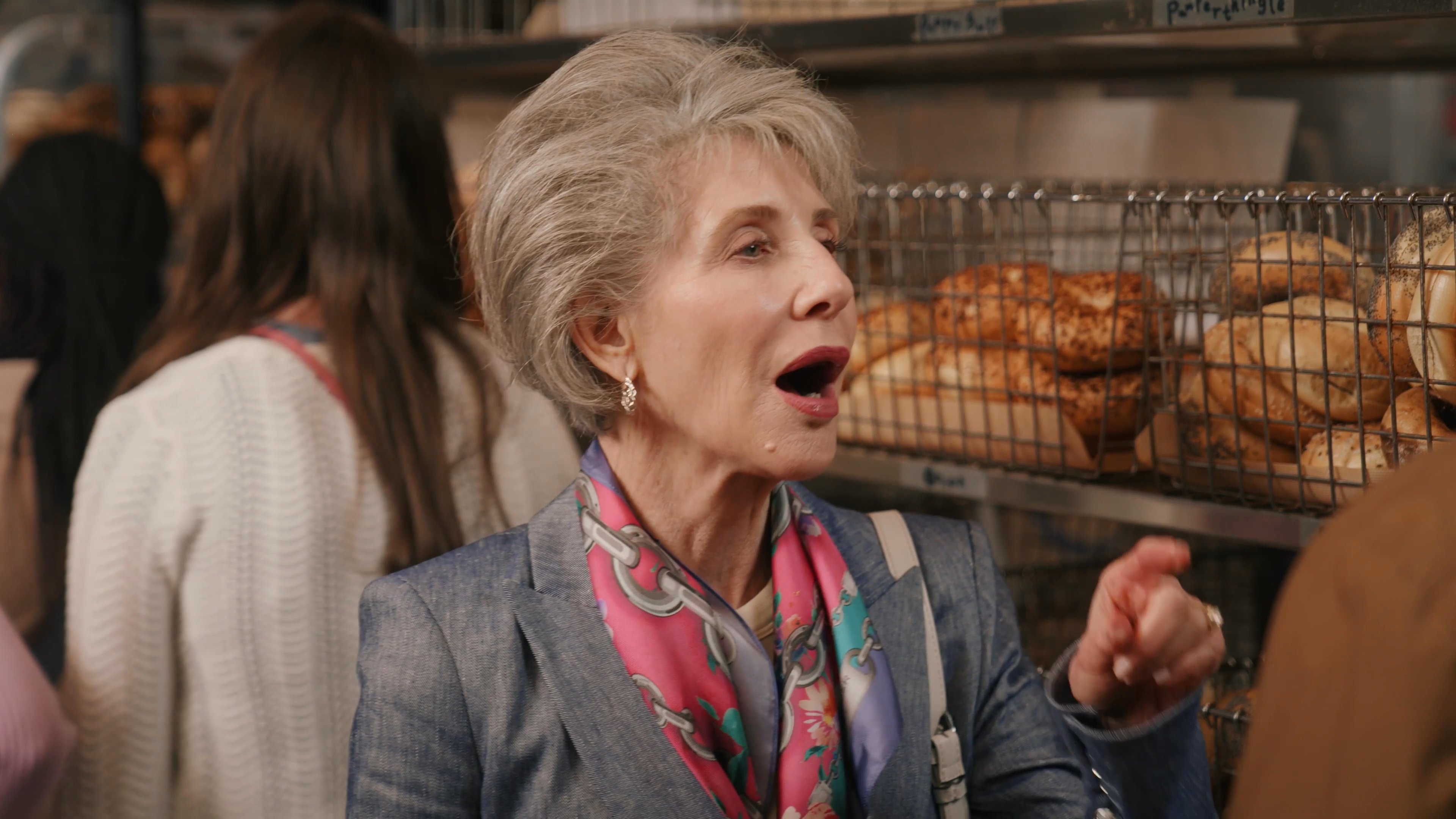 An older woman in a blazer and a younger man engage in a lighthearted conversation inside a busy bakery. The scene focuses on their interpersonal dynamic as they stand before shelves stocked with baked goods.