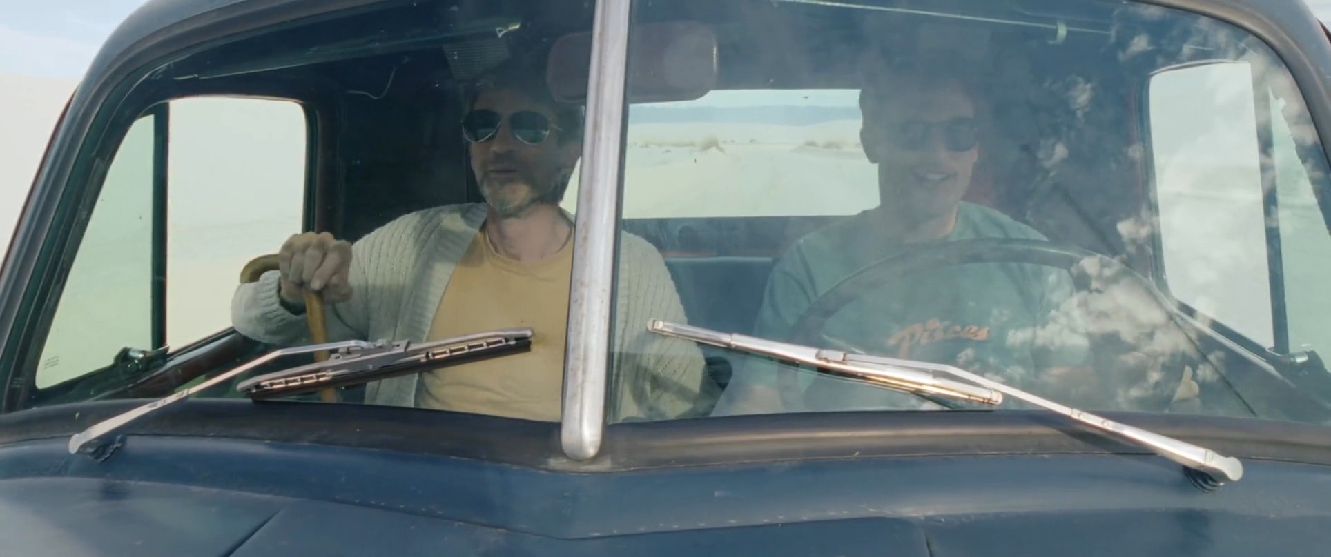 Two individuals travel in a vintage truck along a winding road surrounded by expansive white desert dunes. The scene captures the journey as the vehicle moves through the unique, bright landscape.