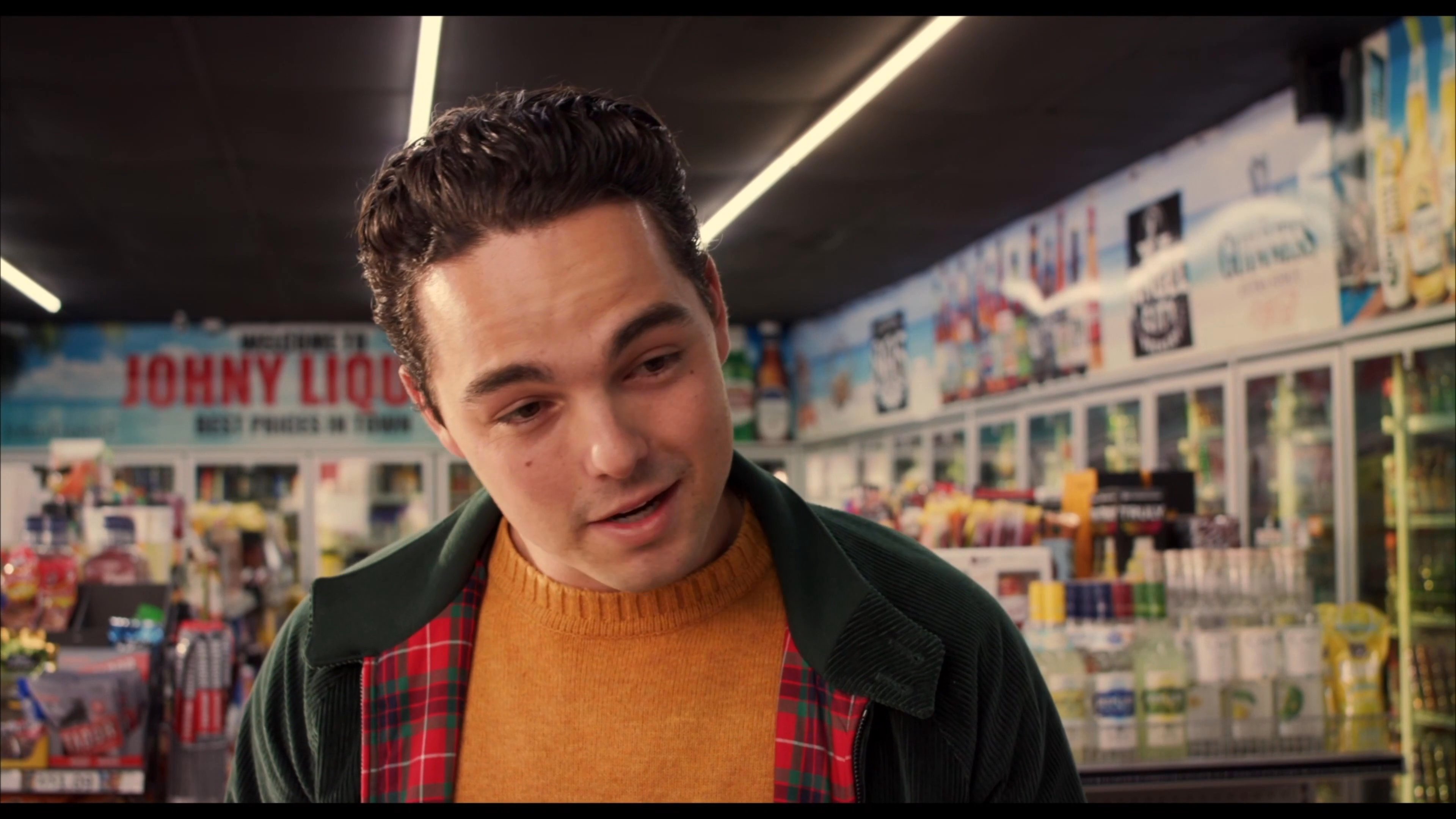 A man speaks directly to the camera while standing in the aisles of a liquor store, gesturing expressively as he talks.