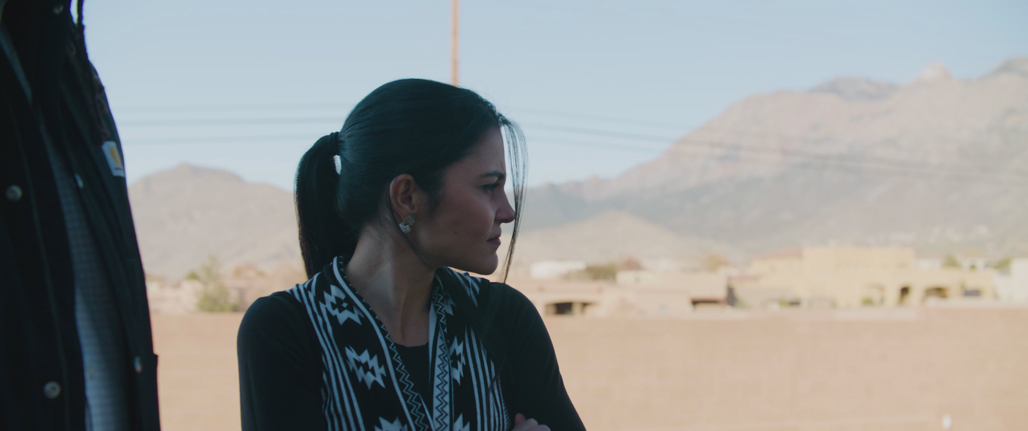 A group of individuals engage in an intense, emotional discussion outdoors in a desert neighborhood. As the conversation concludes, a woman continues the dialogue with another man against the backdrop of an arid mountain landscape.