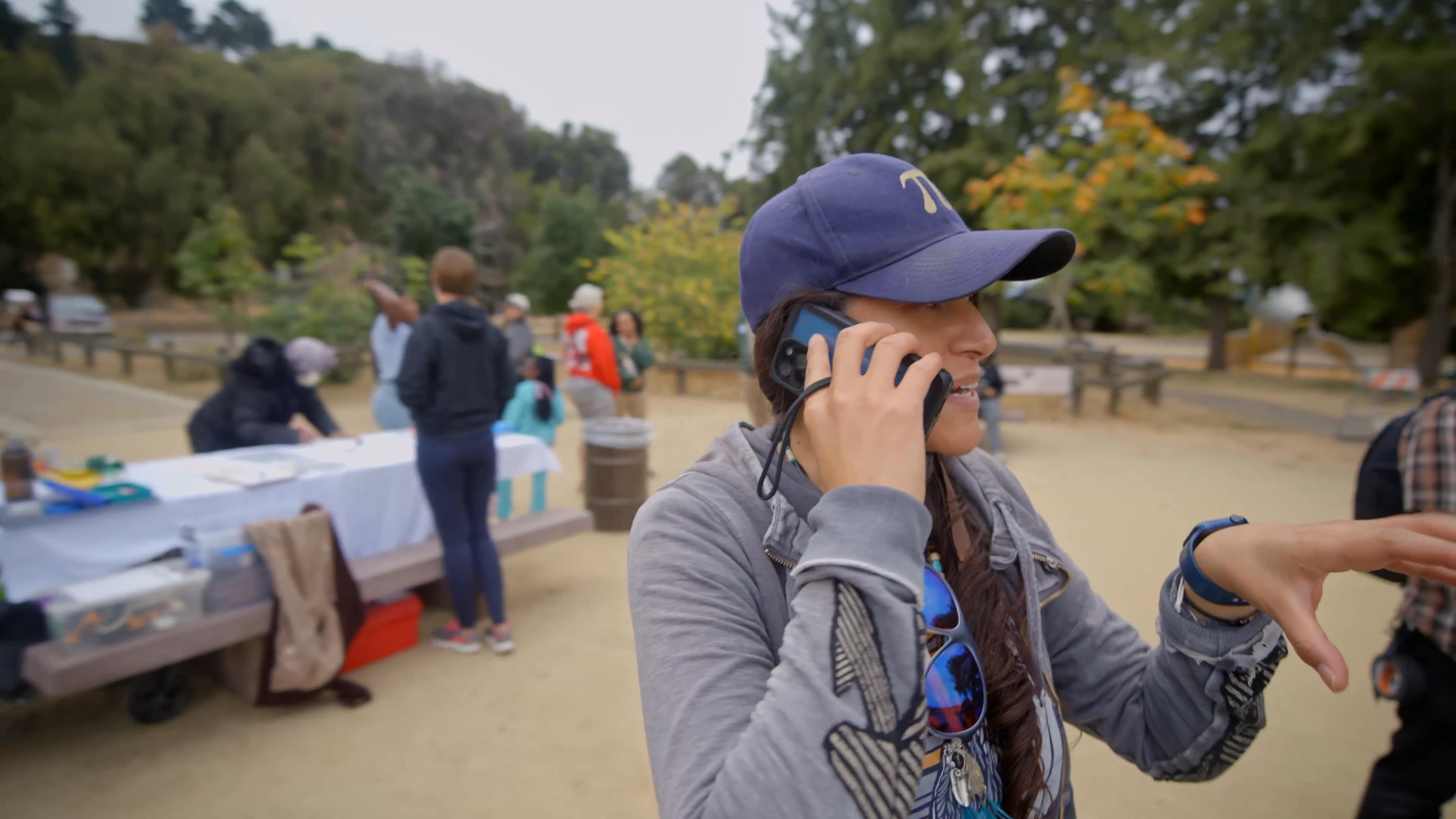 At an outdoor park gathering, a woman in a baseball cap welcomes attendees and explains how she wants everyone to feel valued. She greets a young girl with a warm hug as volunteers and guests gather nearby.