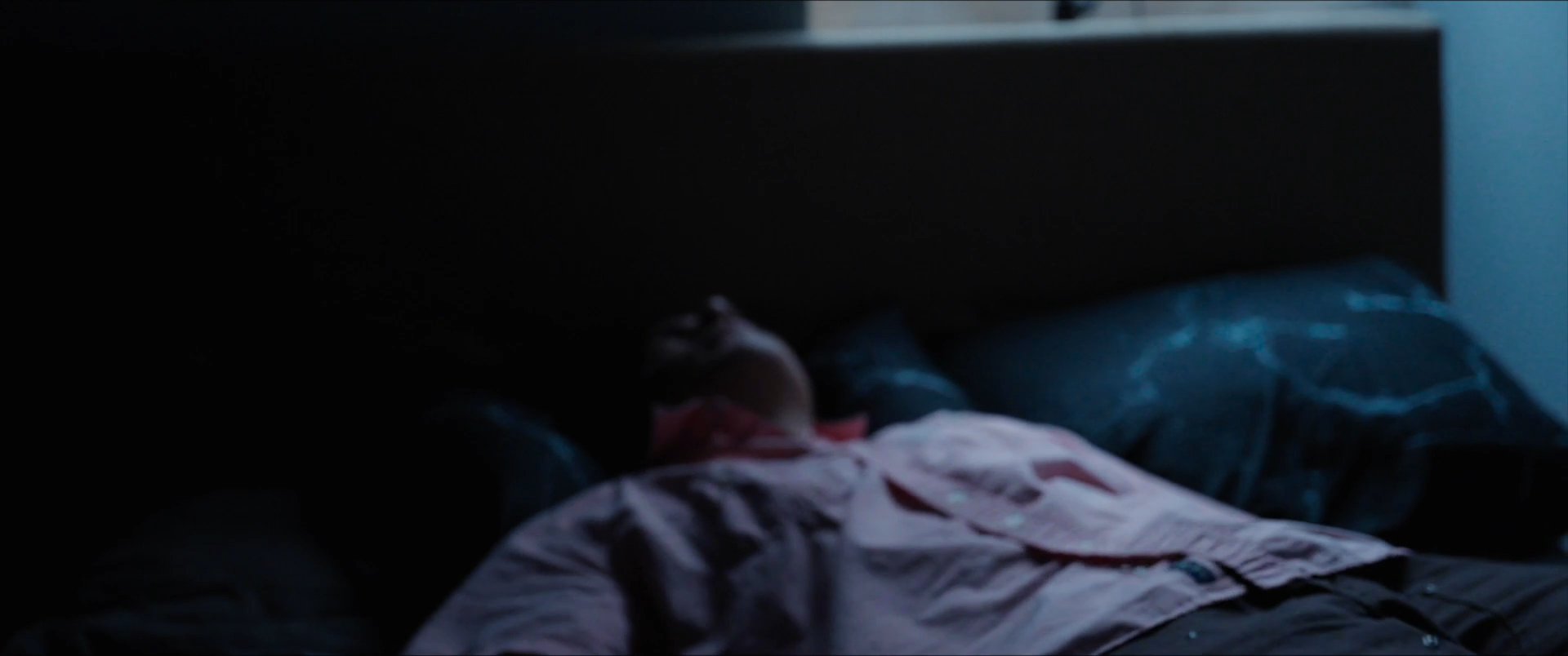 A man in a dimly lit bedroom moves from standing in a doorway to lying down and drifting off to sleep. The sequence emphasizes a quiet, contemplative atmosphere during the late night hours.