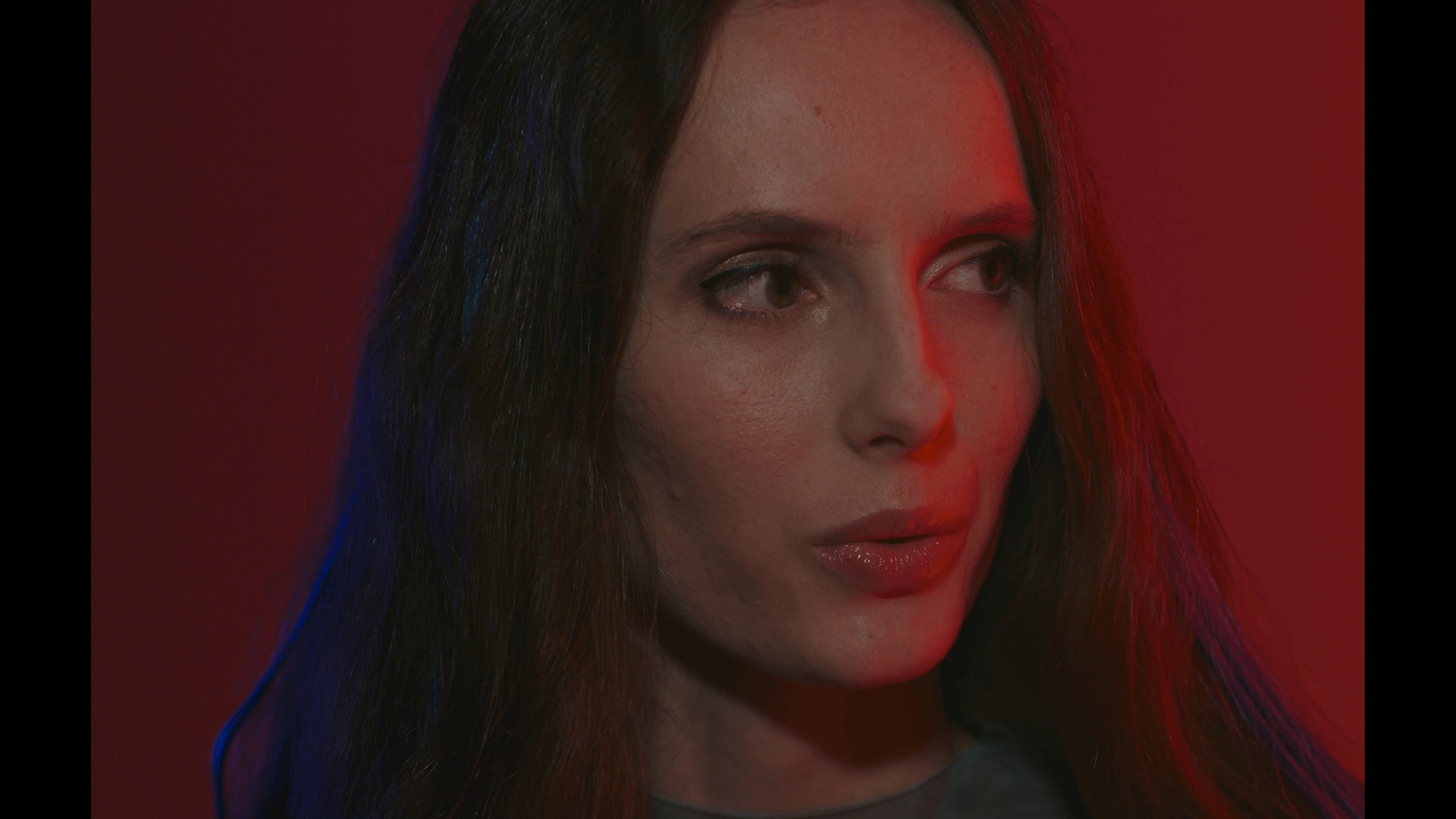 An individual interacts with a floating holographic grid of images while being monitored through a sophisticated facial recognition interface. The scene transitions between high-tech digital projections and a minimalist, red-lit physical environment.