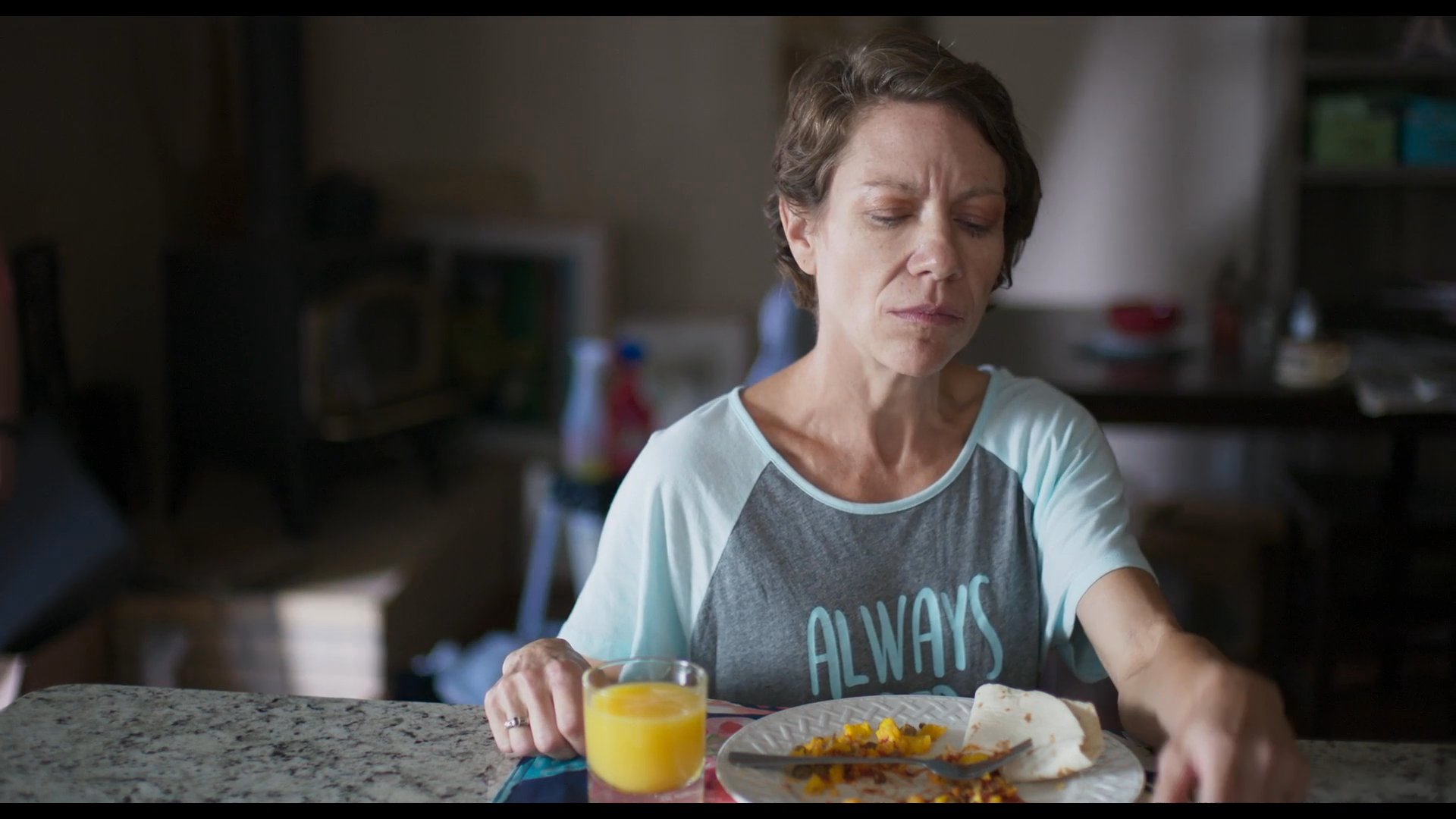 A woman sits at a kitchen table, eating breakfast while appearing absorbed in deep thought. She occasionally touches her face and neck as she reflects during her meal.