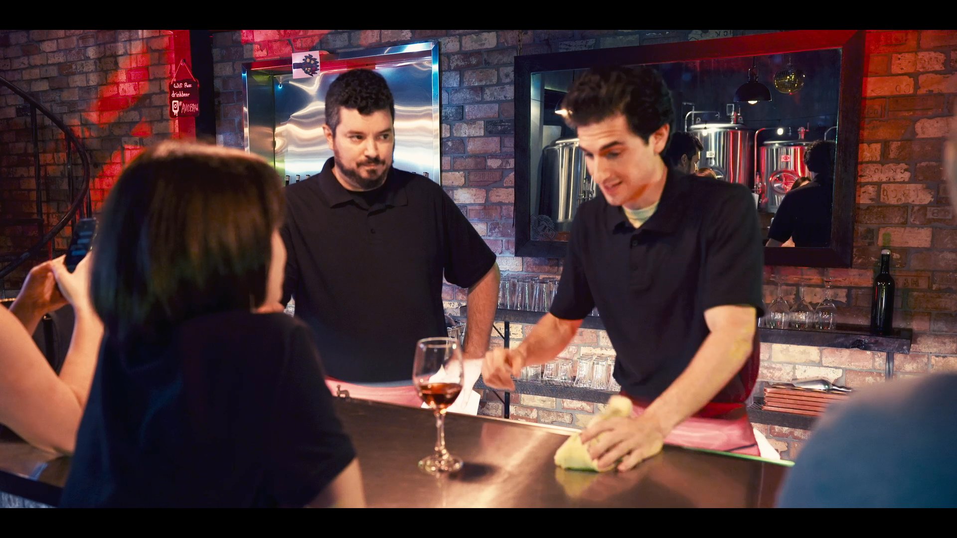 Two bartenders discuss service techniques while cleaning the bar counter at a brewery. A colleague observes the demonstration as they engage with a customer seated at the bar.