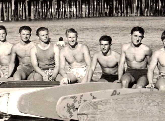 A large group of men sits together on a row of surfboards at the beach, posing for a group portrait in front of a wooden pier.