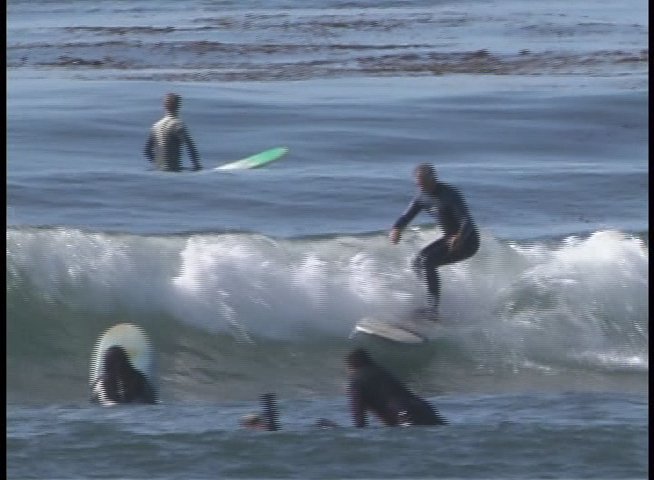 A skilled surfer maneuvers across the face of a rolling ocean wave while others wait in the lineup. The scene captures the fluid motion and technical balance required for competitive surfing.