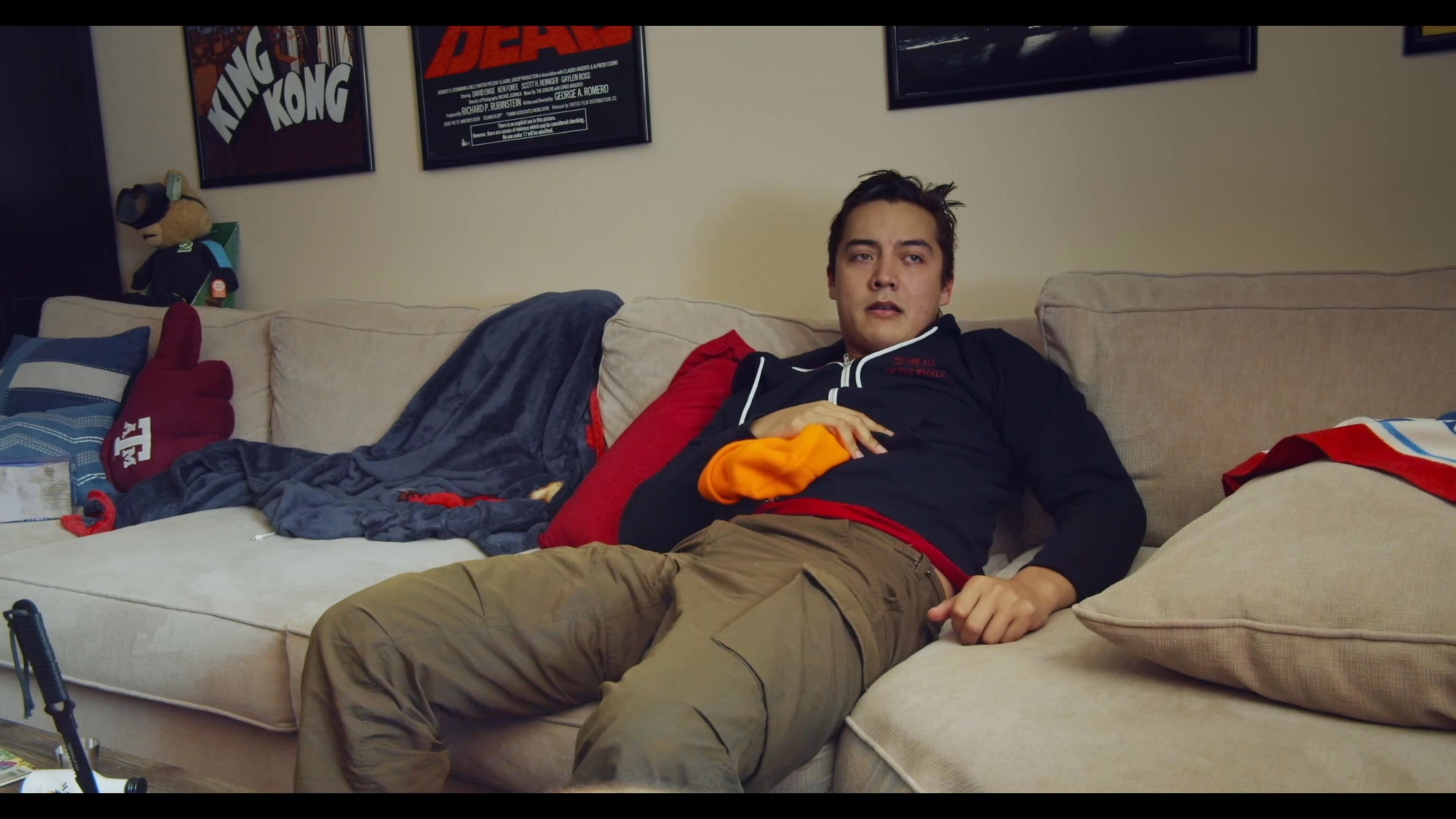 Young men transition between periods of post-party relaxation on a sofa and tidying up a cluttered living room. The scene captures the casual, slow-paced atmosphere of domestic life in an apartment.