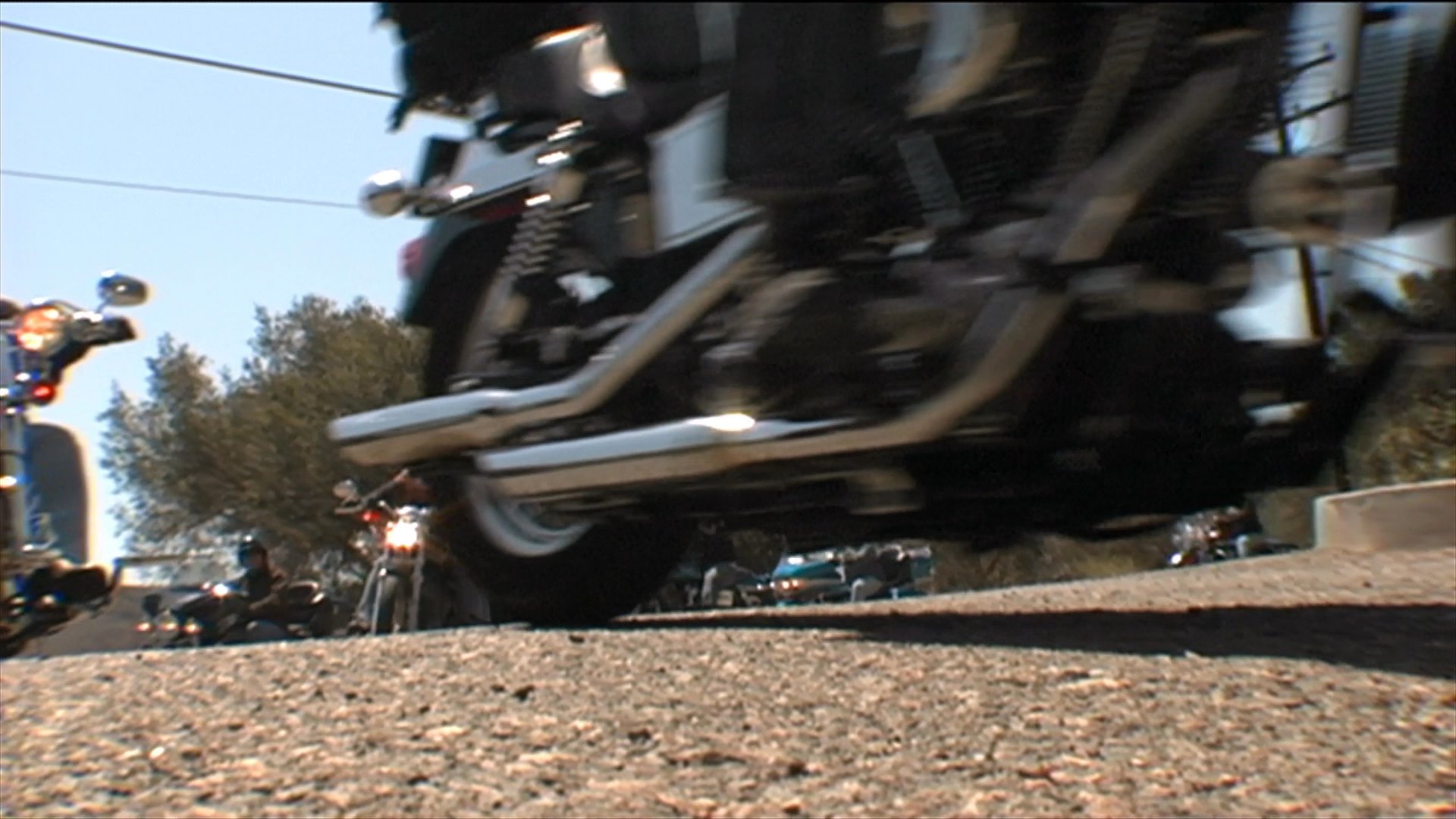 A group of motorcyclists rides in procession along a scenic road in Orange County, California. An individual discusses the group's journey while participants navigate the path on various motorcycles.